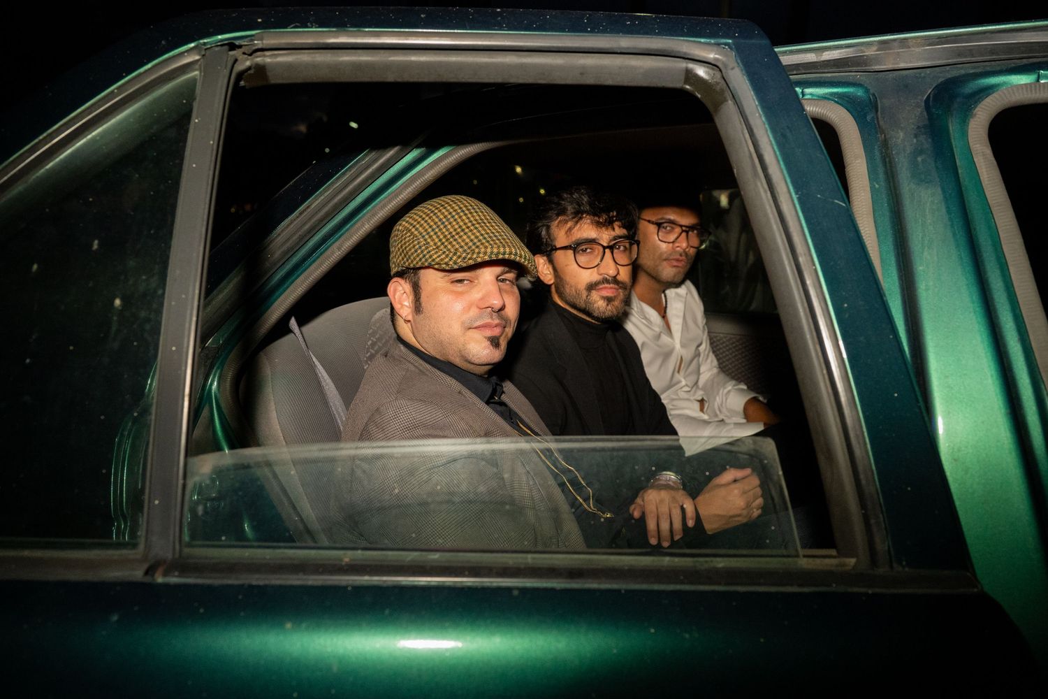 Three men sitting inside a green car photographed through the window at night.