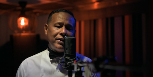 Male vocalist singing into studio microphone in warmly lit professional recording studio.