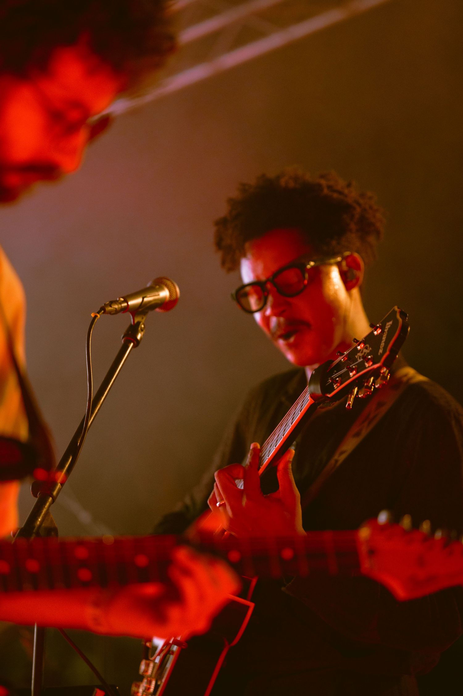 A curly-haired guitarist with glasses playing a red electric guitar on a red-lit stage.