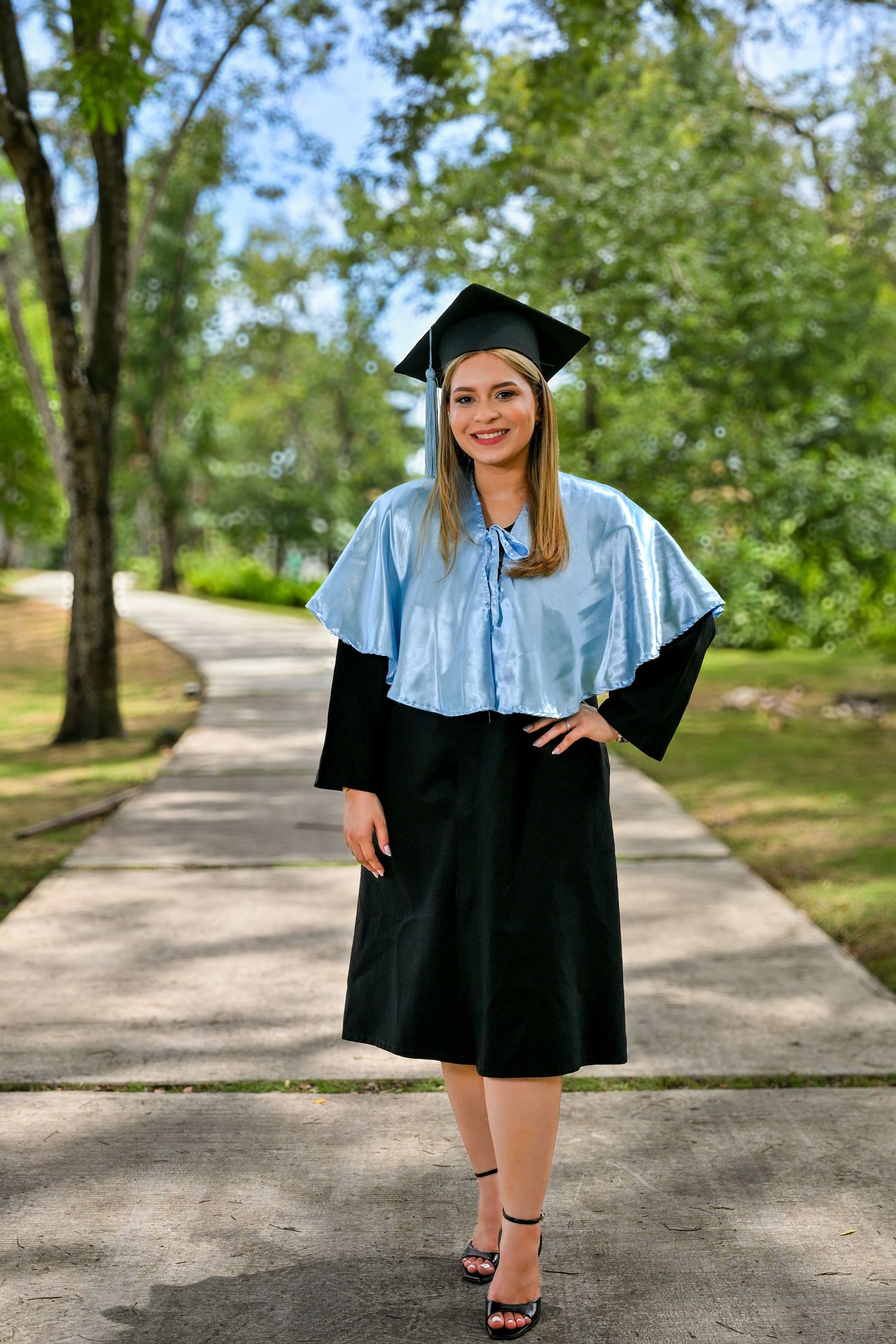 Smiling young woman in black graduation gown with light blue hood and cap posing outdoors on a tree-lined path.