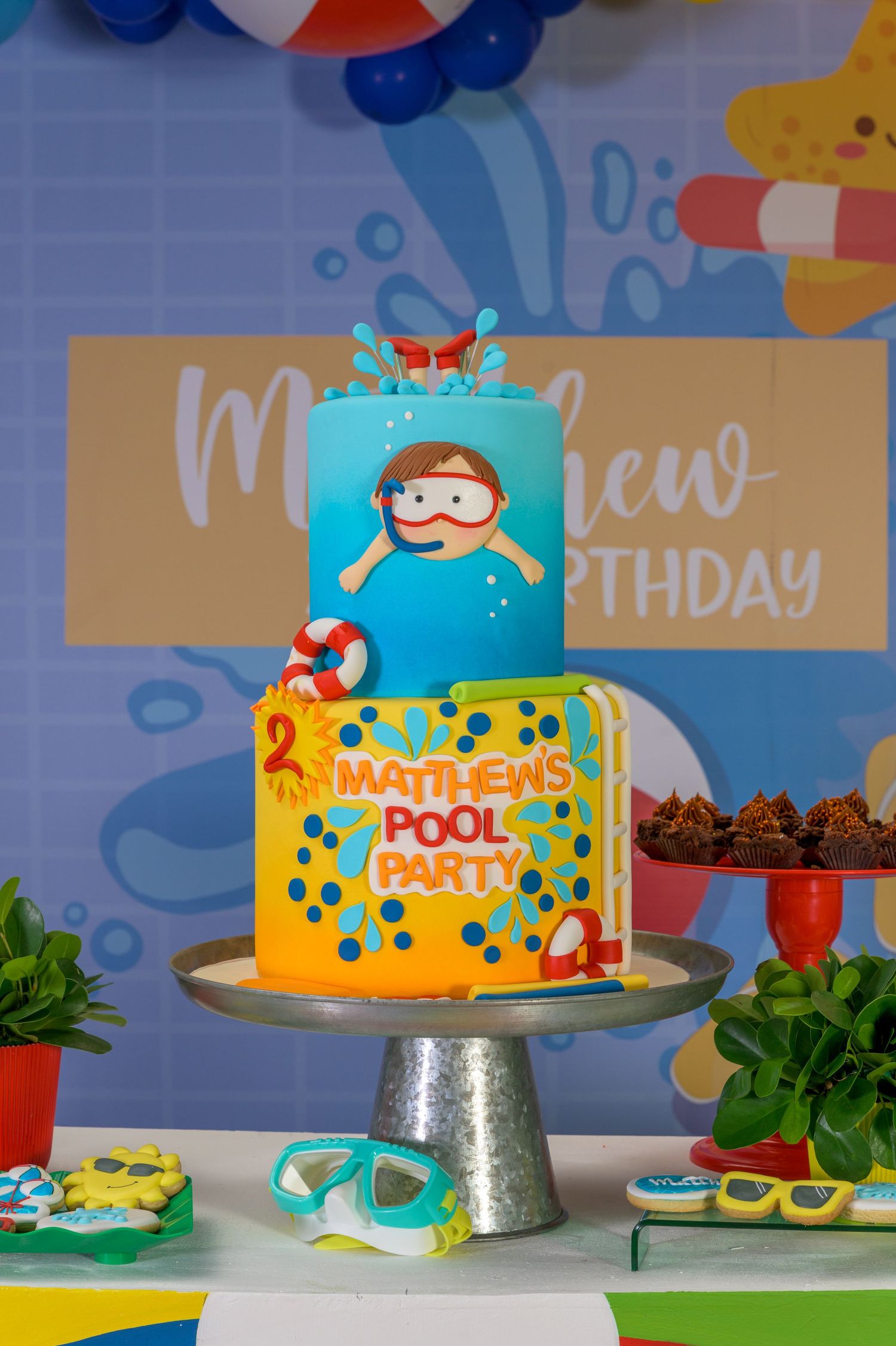 Two-tier Matthews Pool Party birthday cake with swimmer decoration on cake stand at party display.