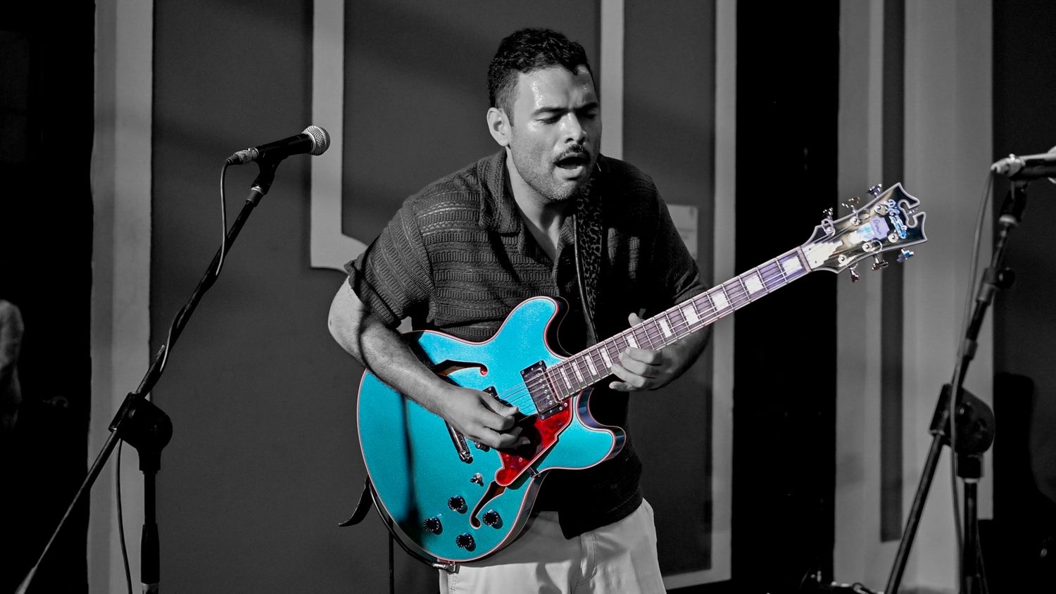 Guitarist playing a teal semi-hollow electric guitar in a selective color photograph.