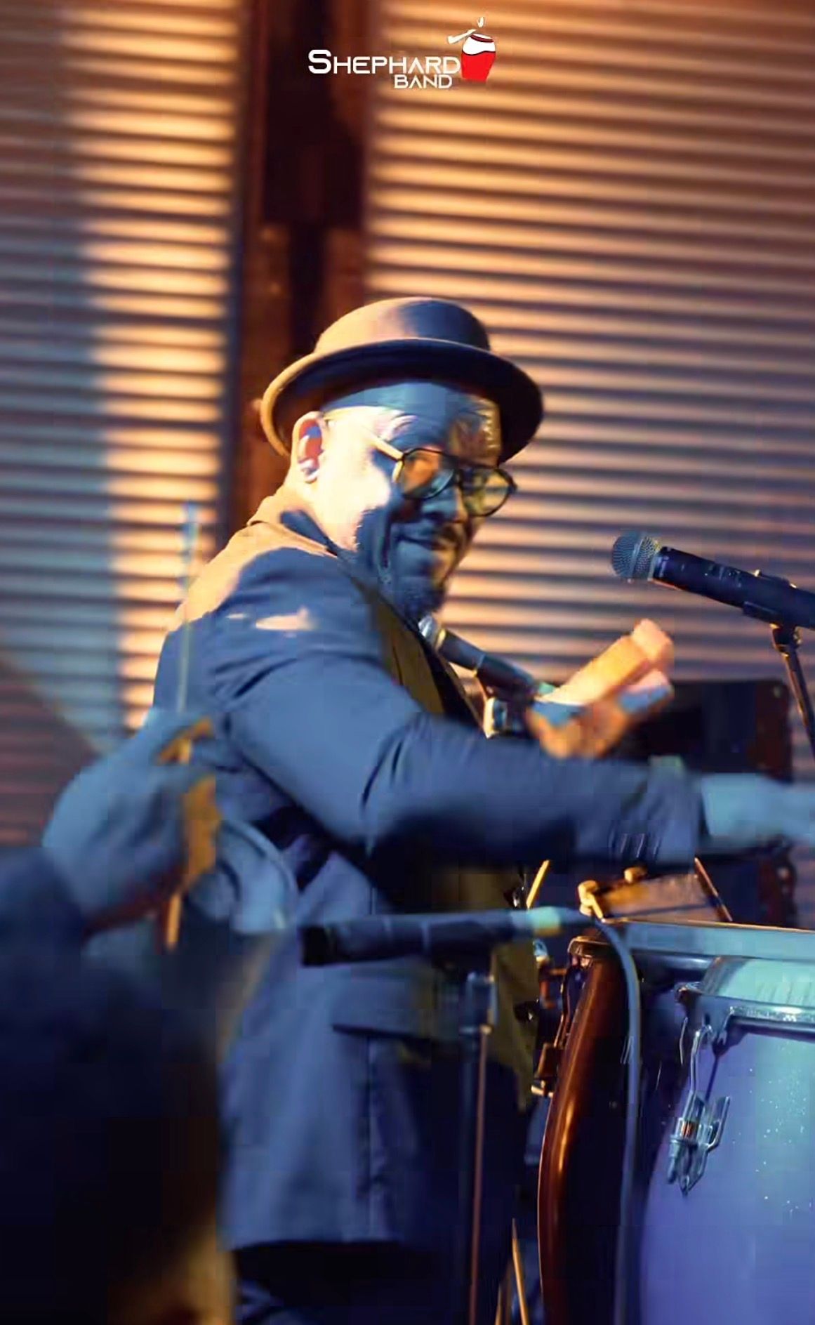 A musician in a bowler hat and suit plays congas on stage under warm dramatic lighting at a live show.