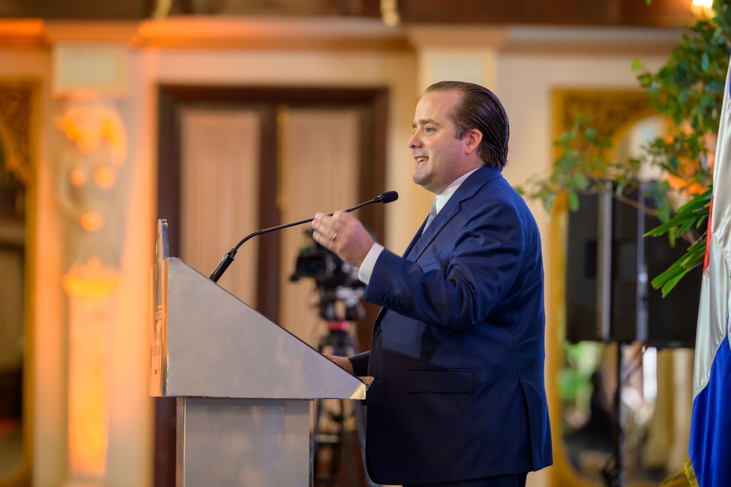 Man in navy suit delivering speech at podium with microphone at formal indoor event.