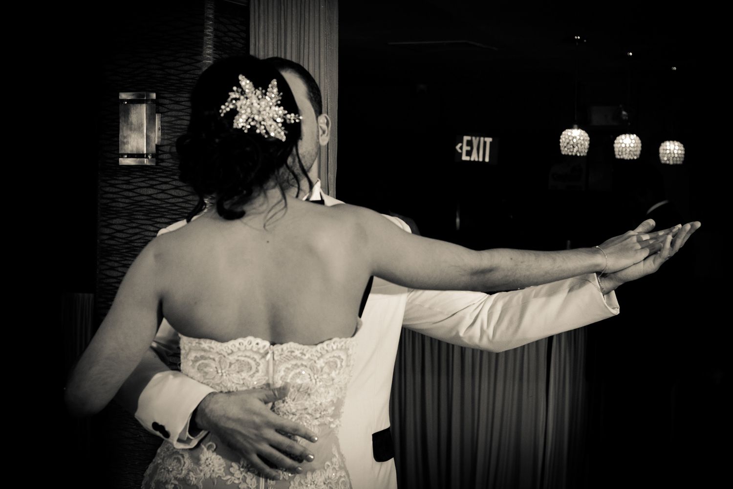 Black and white photo of bride and groom sharing first dance at wedding reception near exit sign.