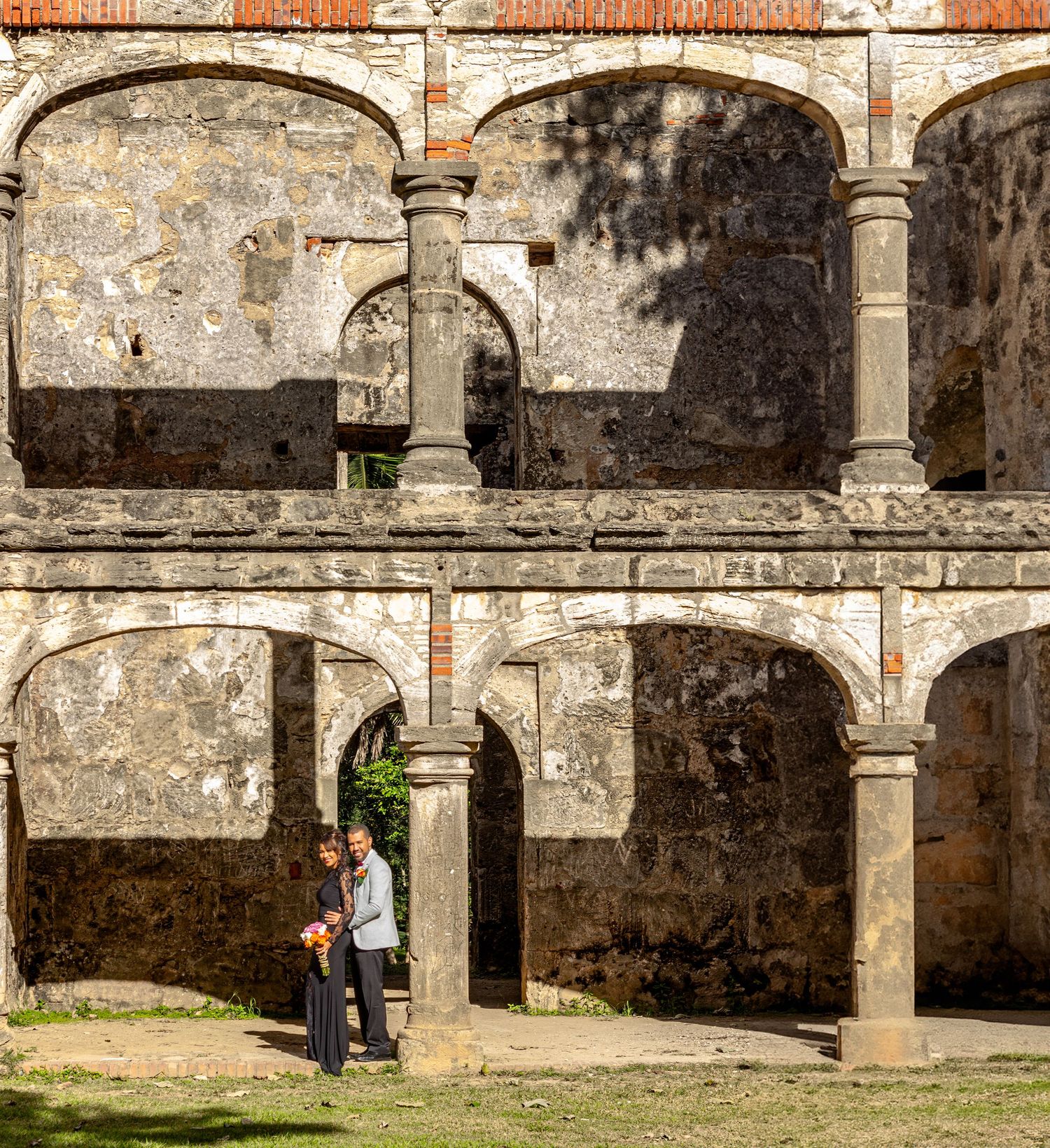 Wedding couple standing under stone arches of historic ruins with bouquet.