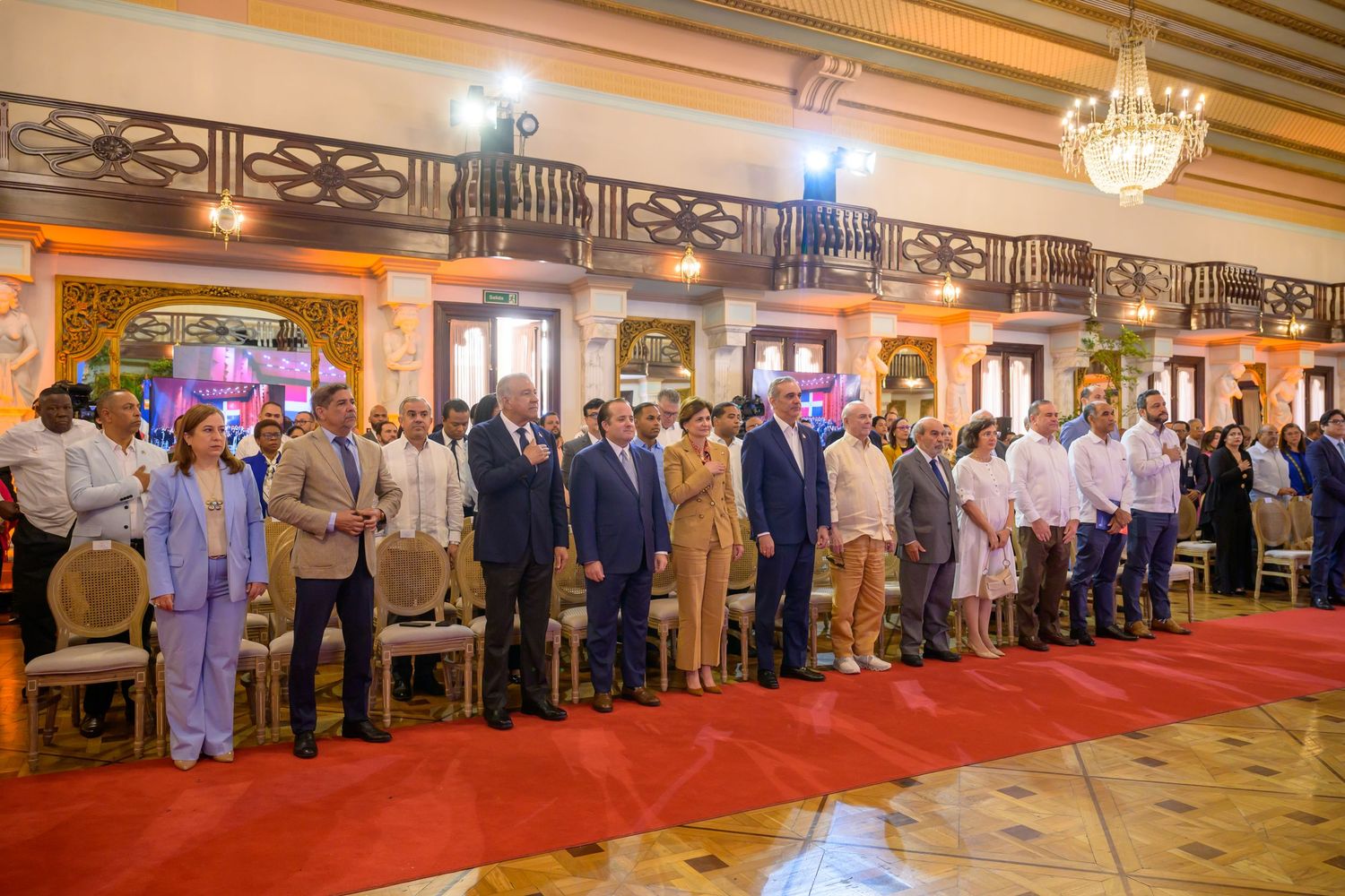 Large group of formally dressed attendees standing together in elegant historic hall.