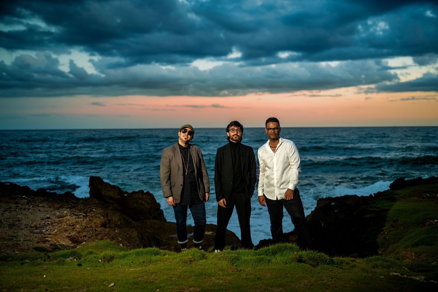 Three men standing on rocky coastal cliffs at sunset with dramatic stormy skies and ocean waves.