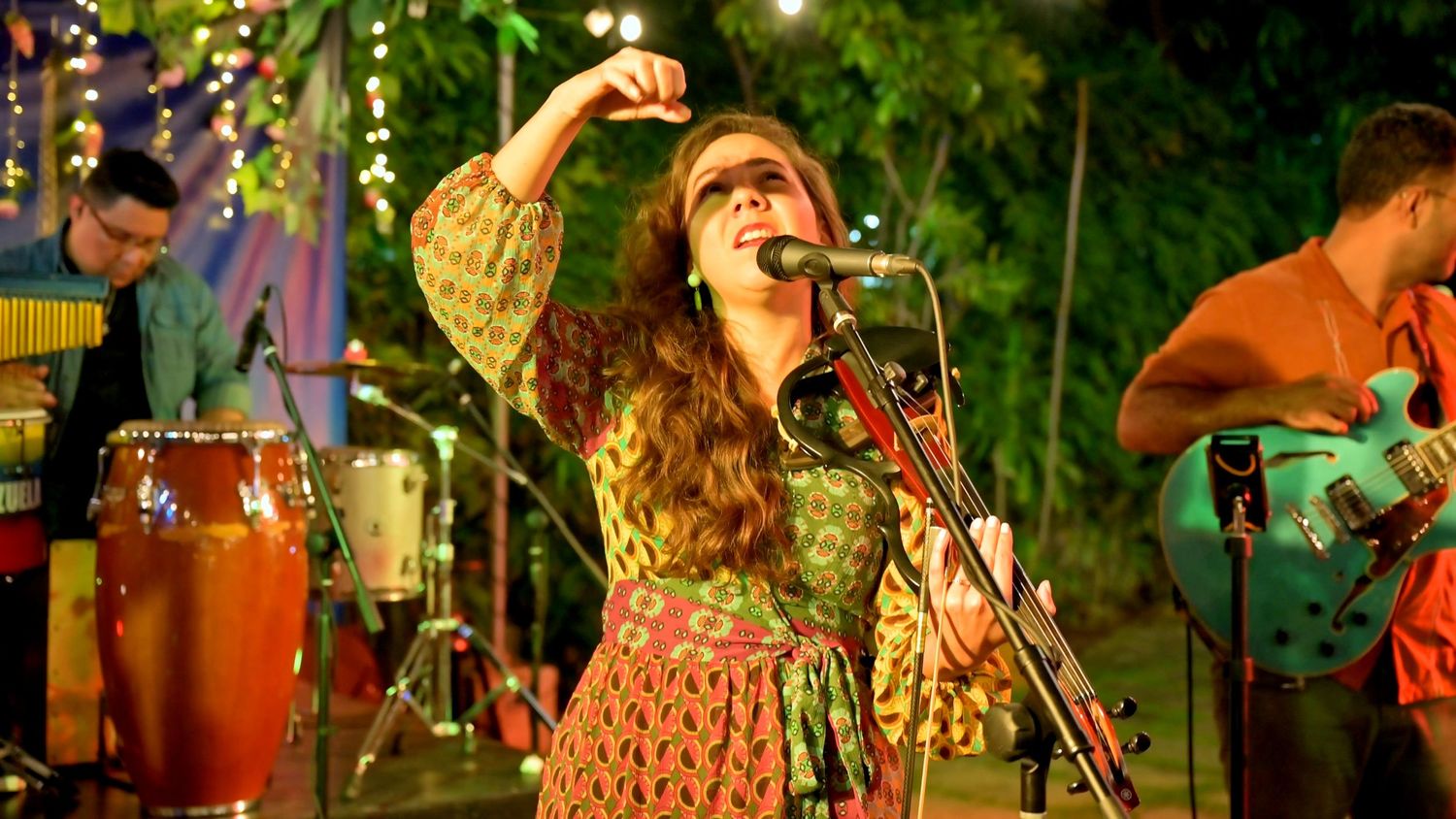 A female singer in a floral dress performs passionately at an outdoor concert with band members visible behind her.