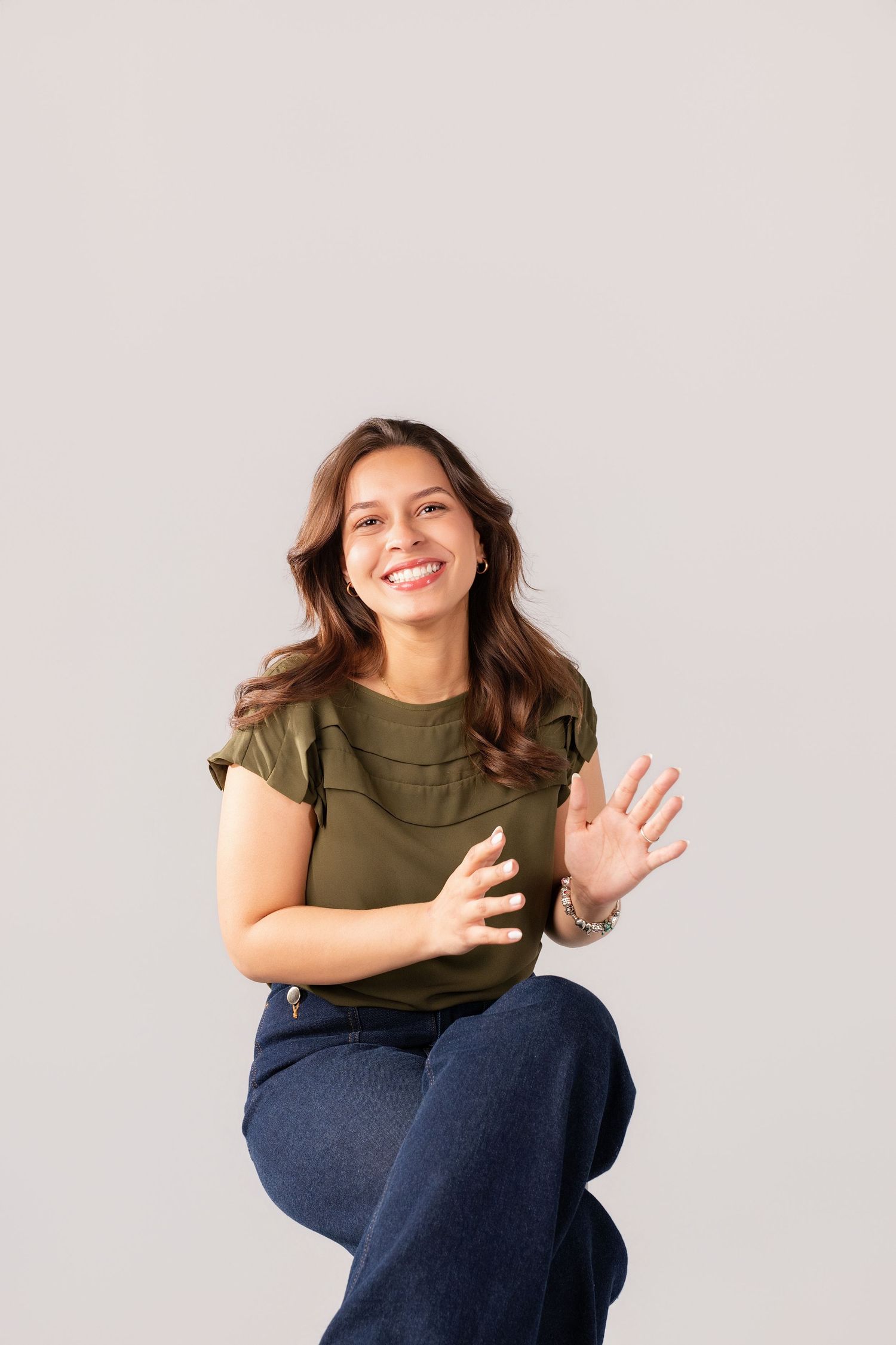 A smiling young woman in a green top and jeans poses casually against a light gray studio background.