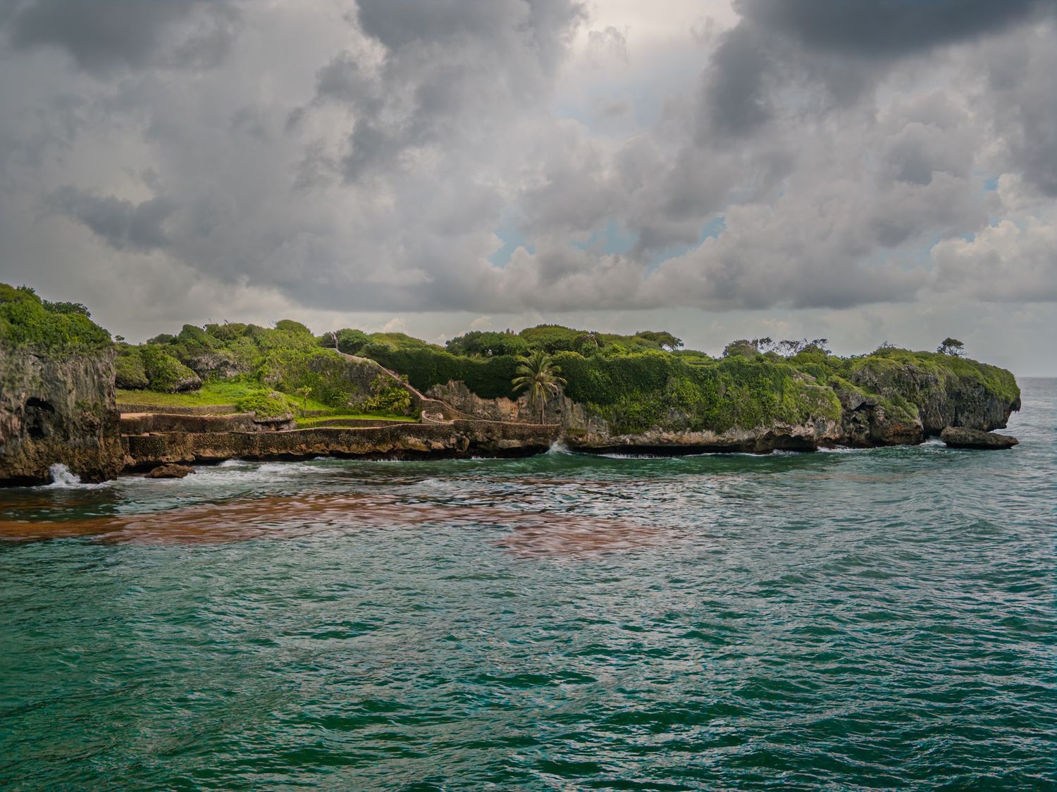 Rocky green island coastline surrounded by choppy turquoise ocean waters under a dramatic overcast sky.
