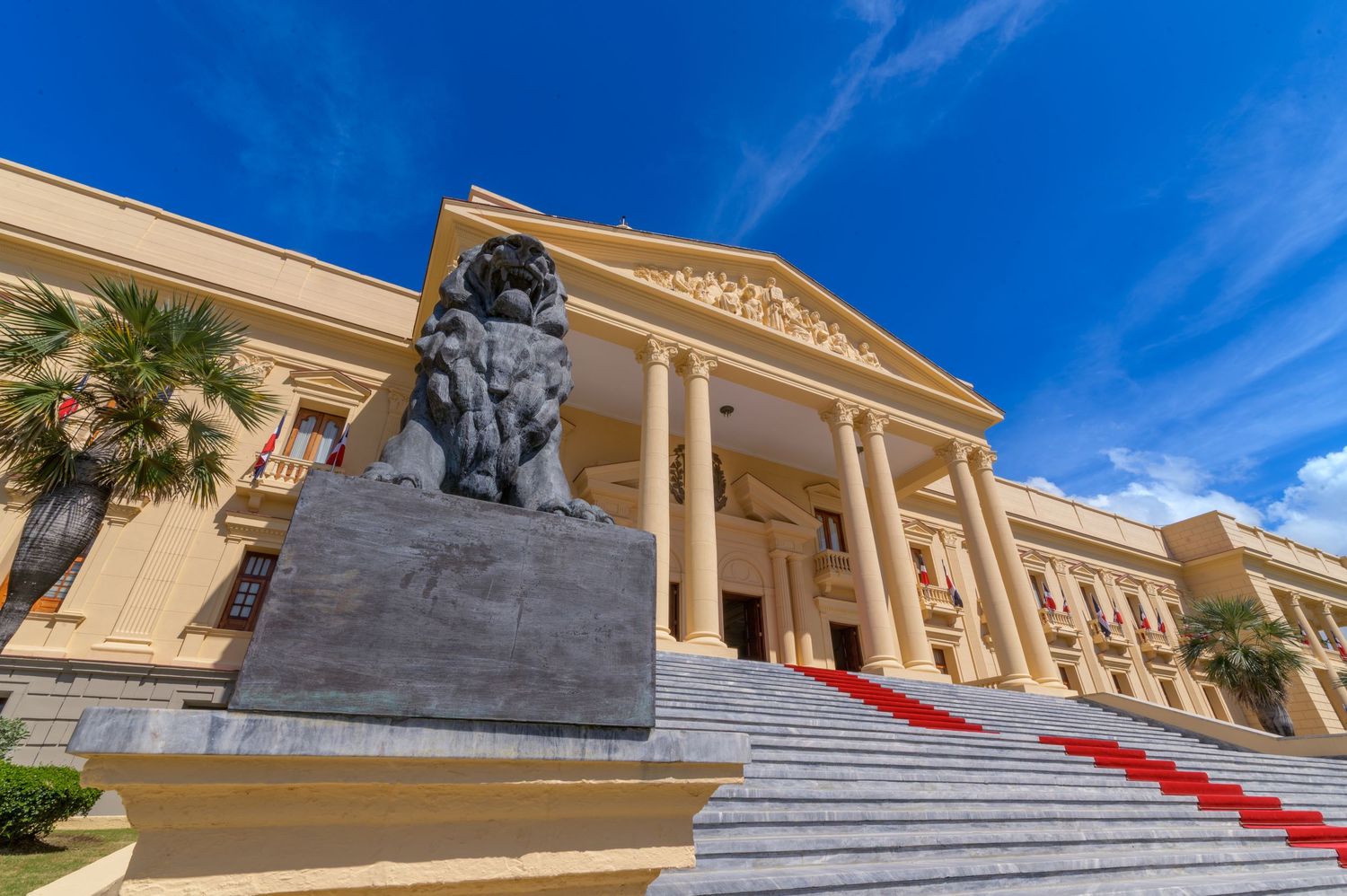 Stone lion statue guards the grand entrance staircase of a neoclassical building with red carpet and tall columns.