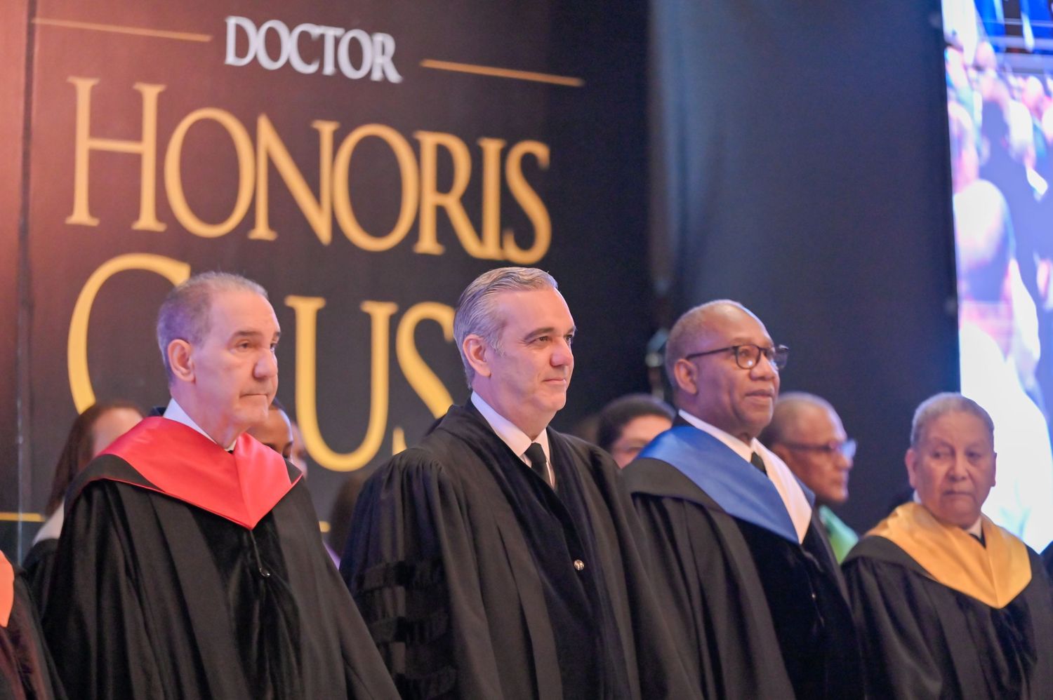 Group of academics in graduation robes standing beneath Doctor Honoris Causa sign at ceremony.