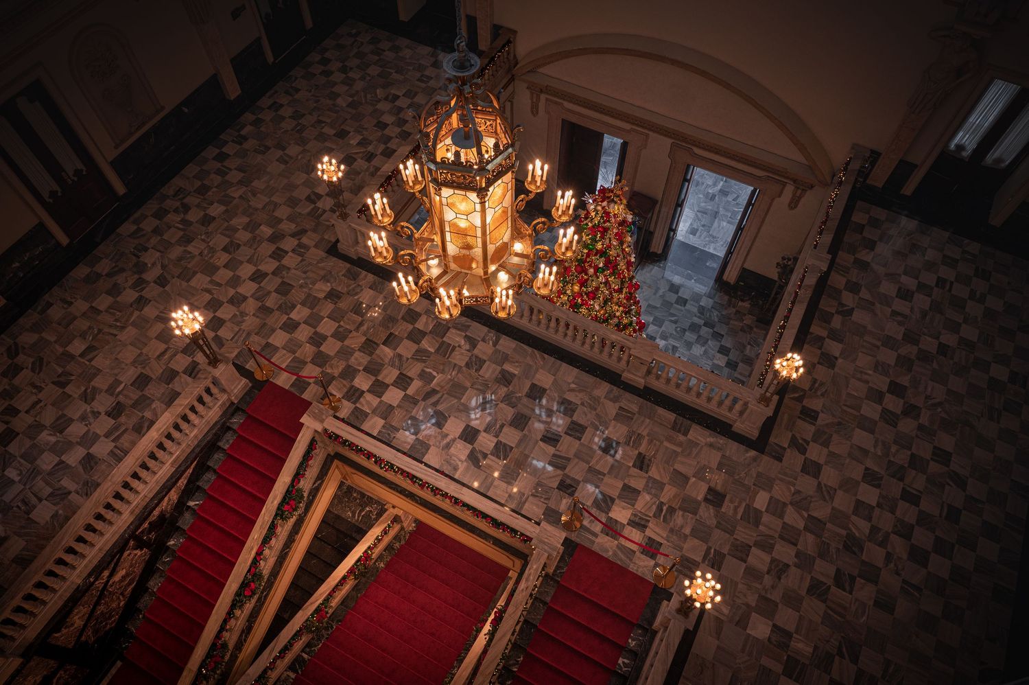 Overhead view of an ornate grand staircase with glowing lantern chandelier and red carpet in a historic building.