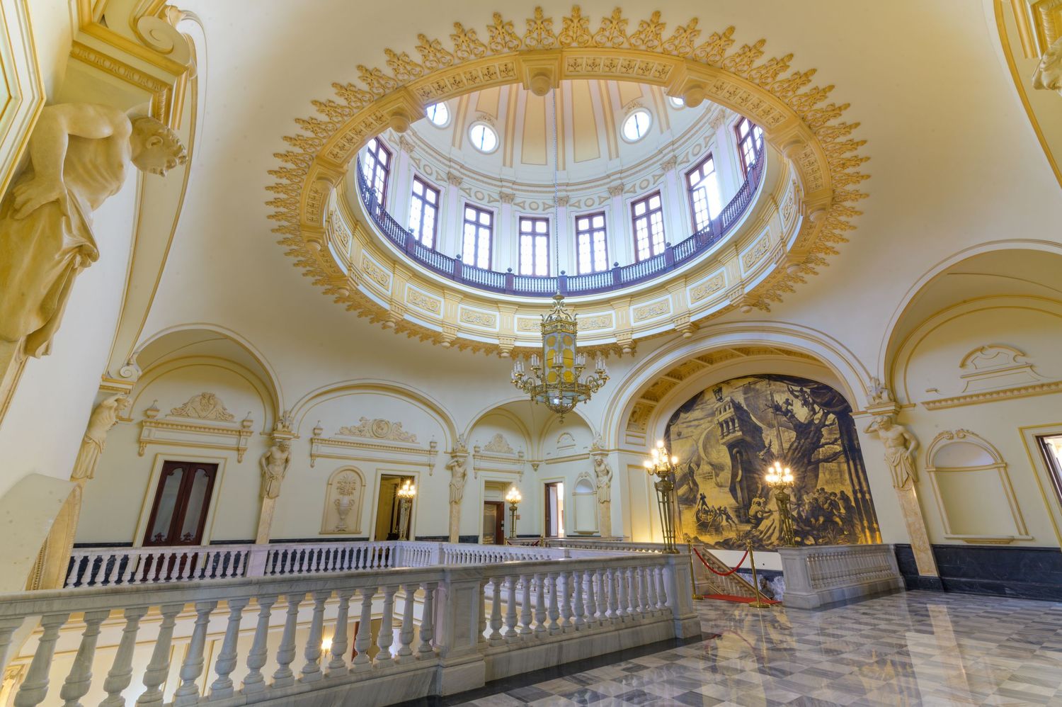 Stunning neoclassical rotunda with gold ornamental ceiling, grand chandelier, marble balustrade, and large wall mural.