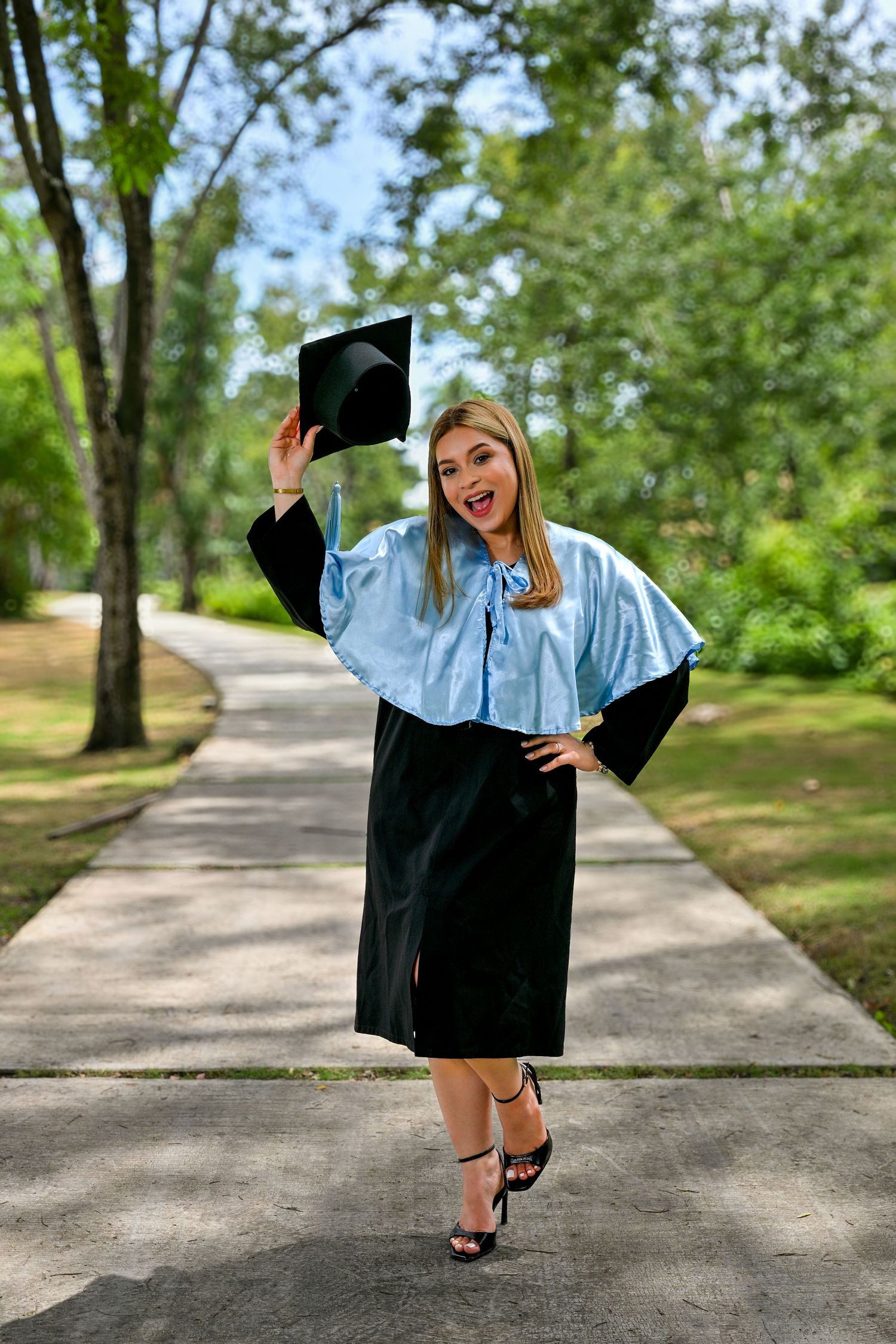 Excited graduate in cap and gown celebrating on a tree-lined pathway outdoors.