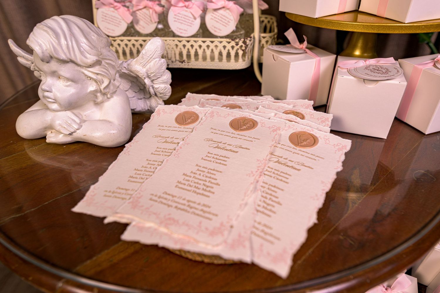 Pink baptism program cards displayed beside an angel figurine and gift boxes.