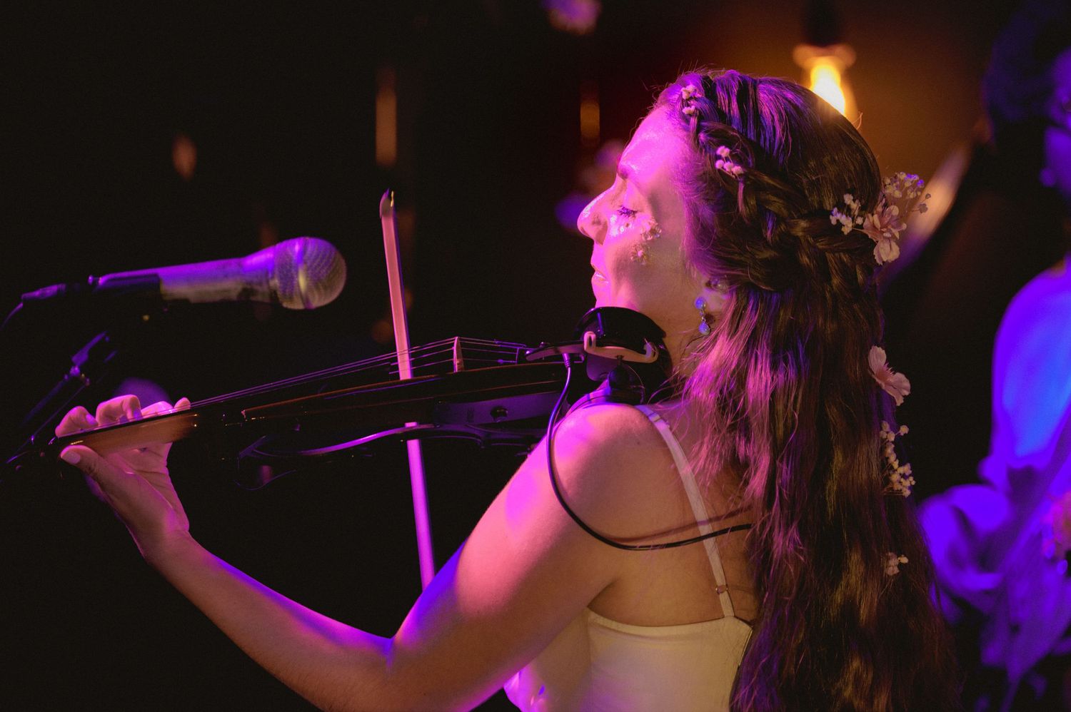 Young woman with floral crown playing electric violin on stage under warm purple stage lighting.