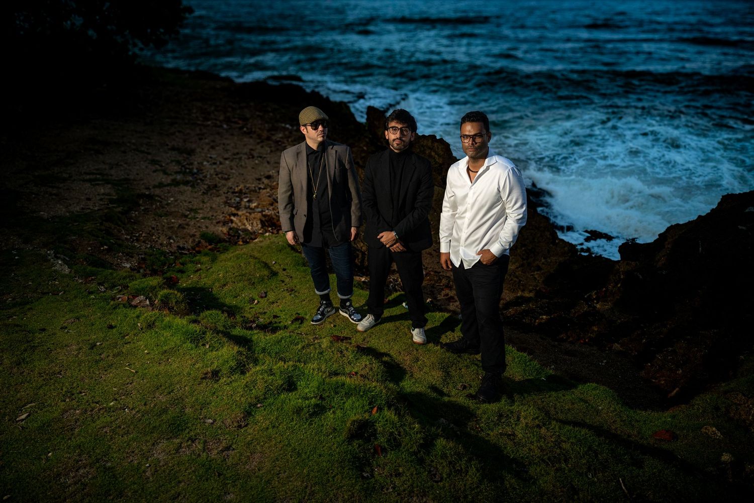 Three men posing on moss-covered rocks by the ocean under moody overcast skies at dusk.
