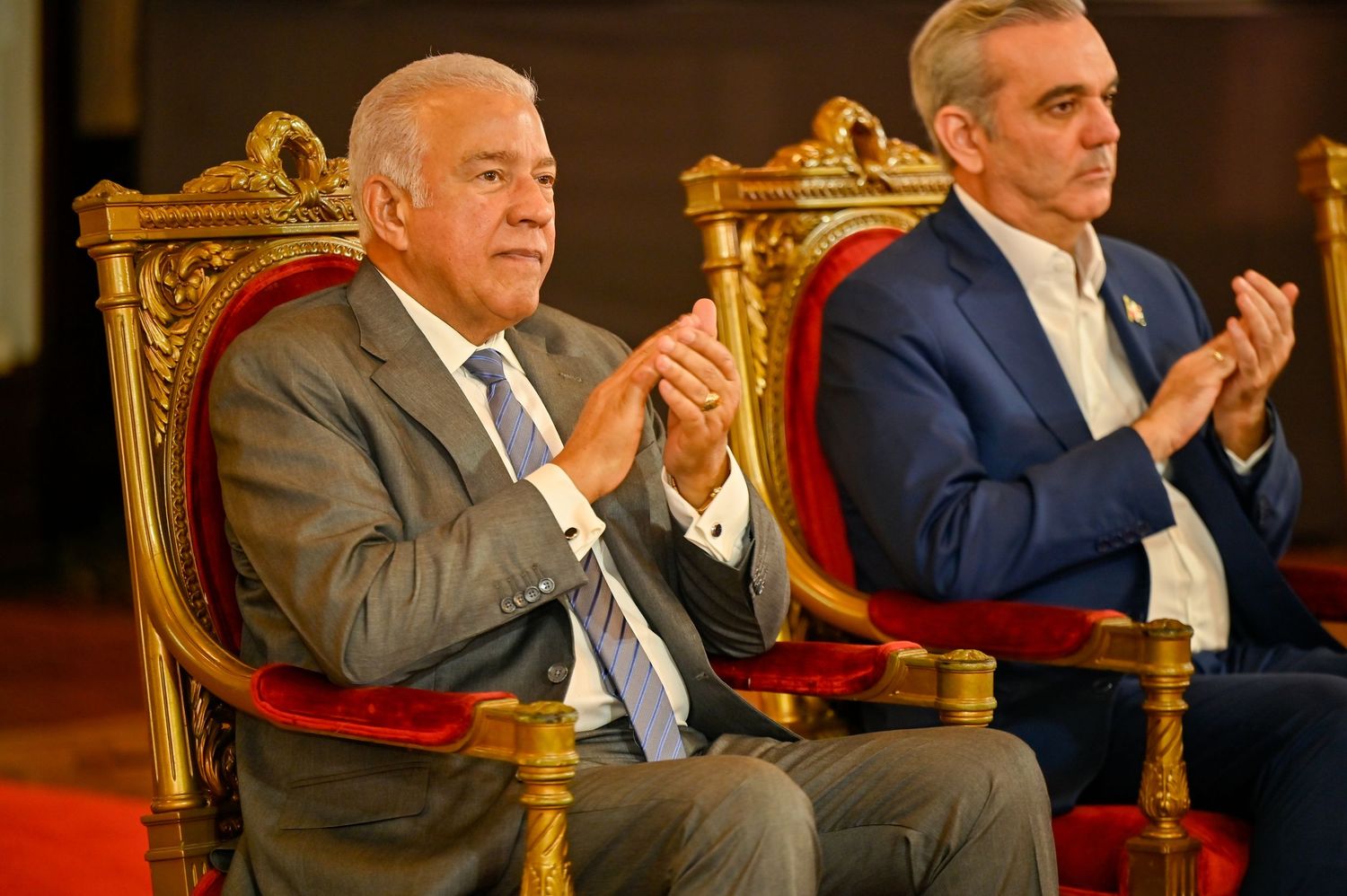 Two men in suits applauding while seated on red velvet ornate chairs at formal ceremony.