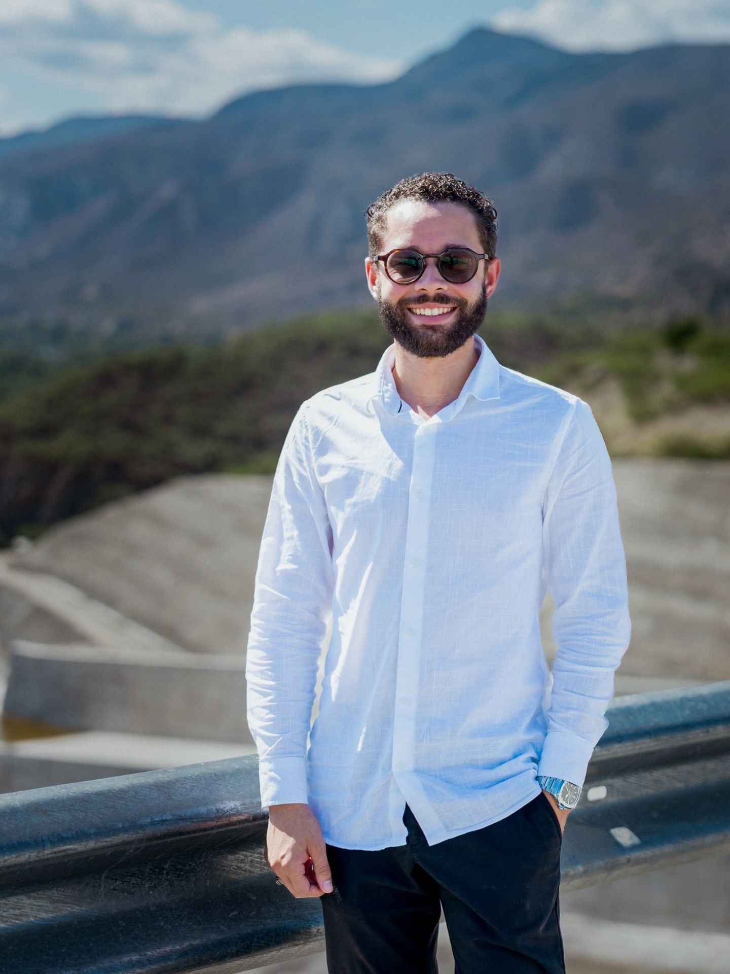 Smiling bearded man in white shirt stands on a bridge with scenic mountain backdrop.