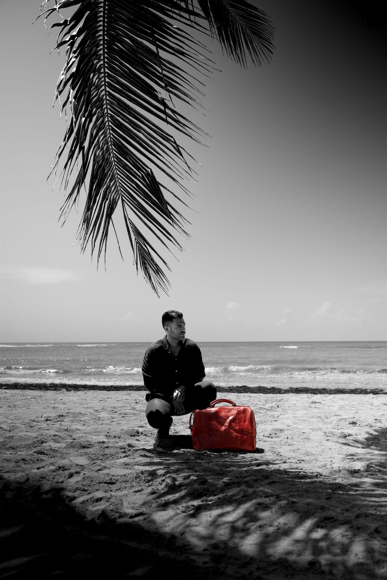 Man sitting on sandy beach beside red bag under palm tree in selective color black and white photo.