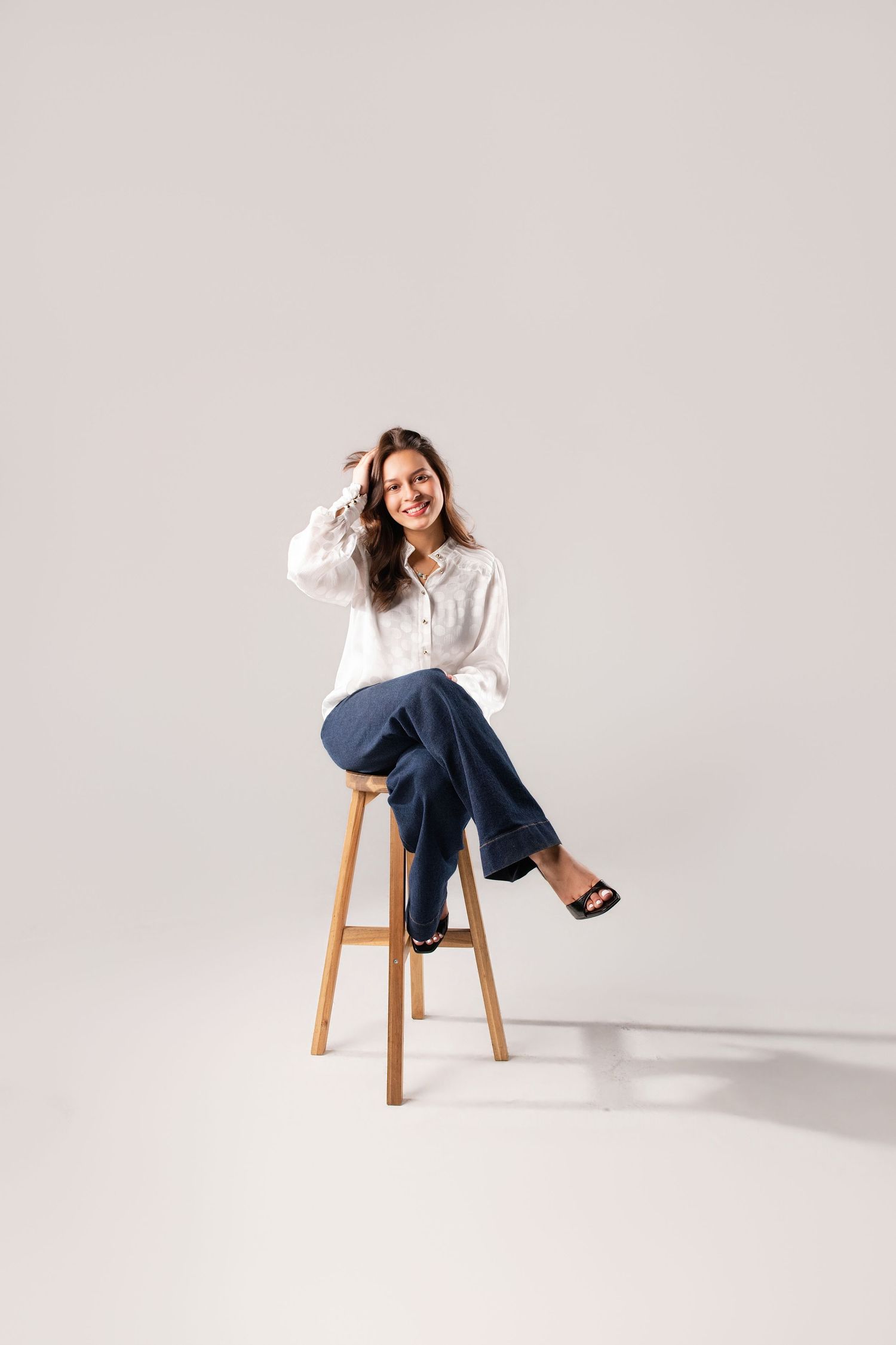 Happy woman in white shirt and blue jeans sitting on wooden stool in studio.