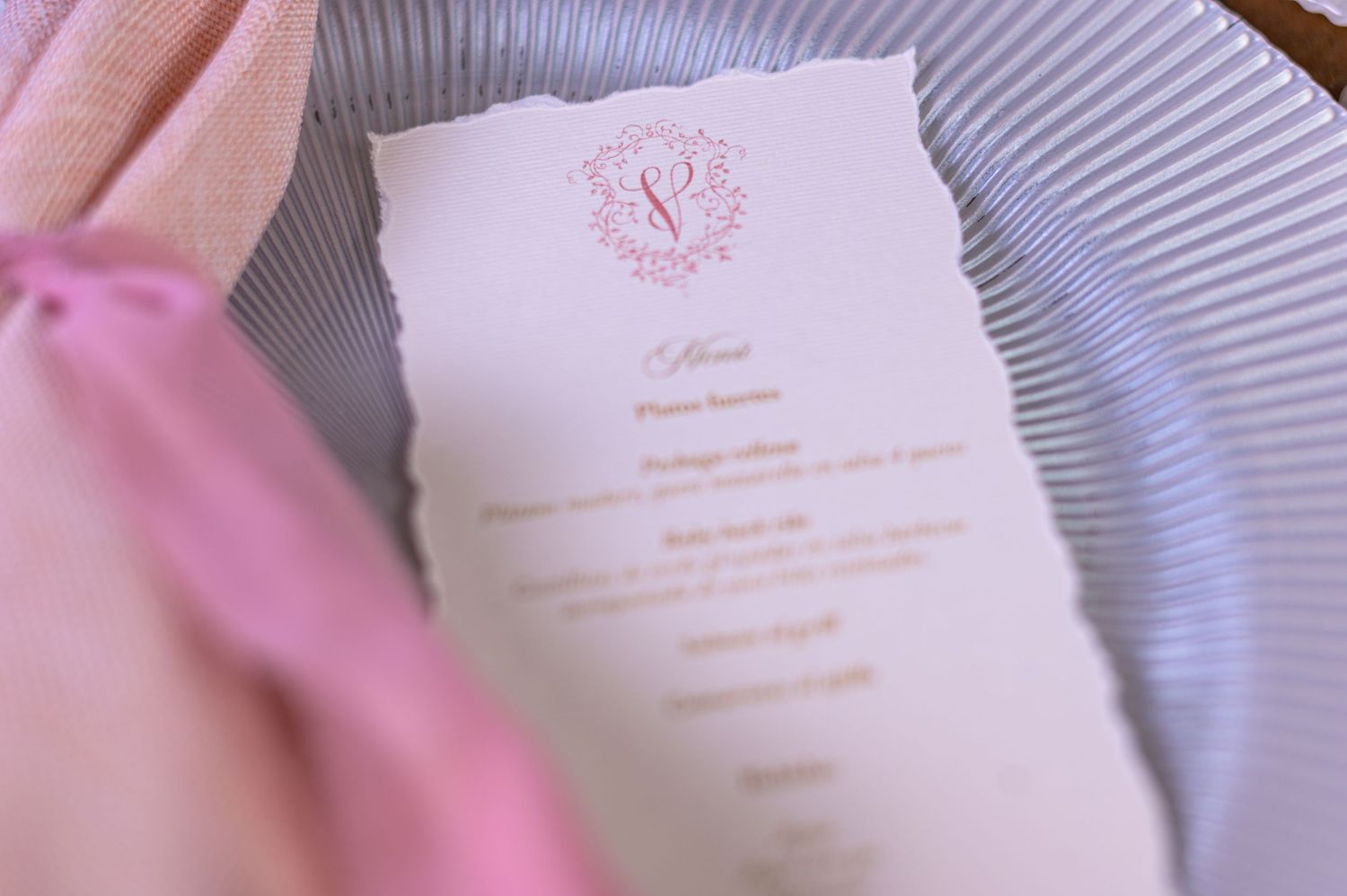 Close-up of an elegant wedding menu card with pink monogram crest on a striped plate with pink ribbon napkin.