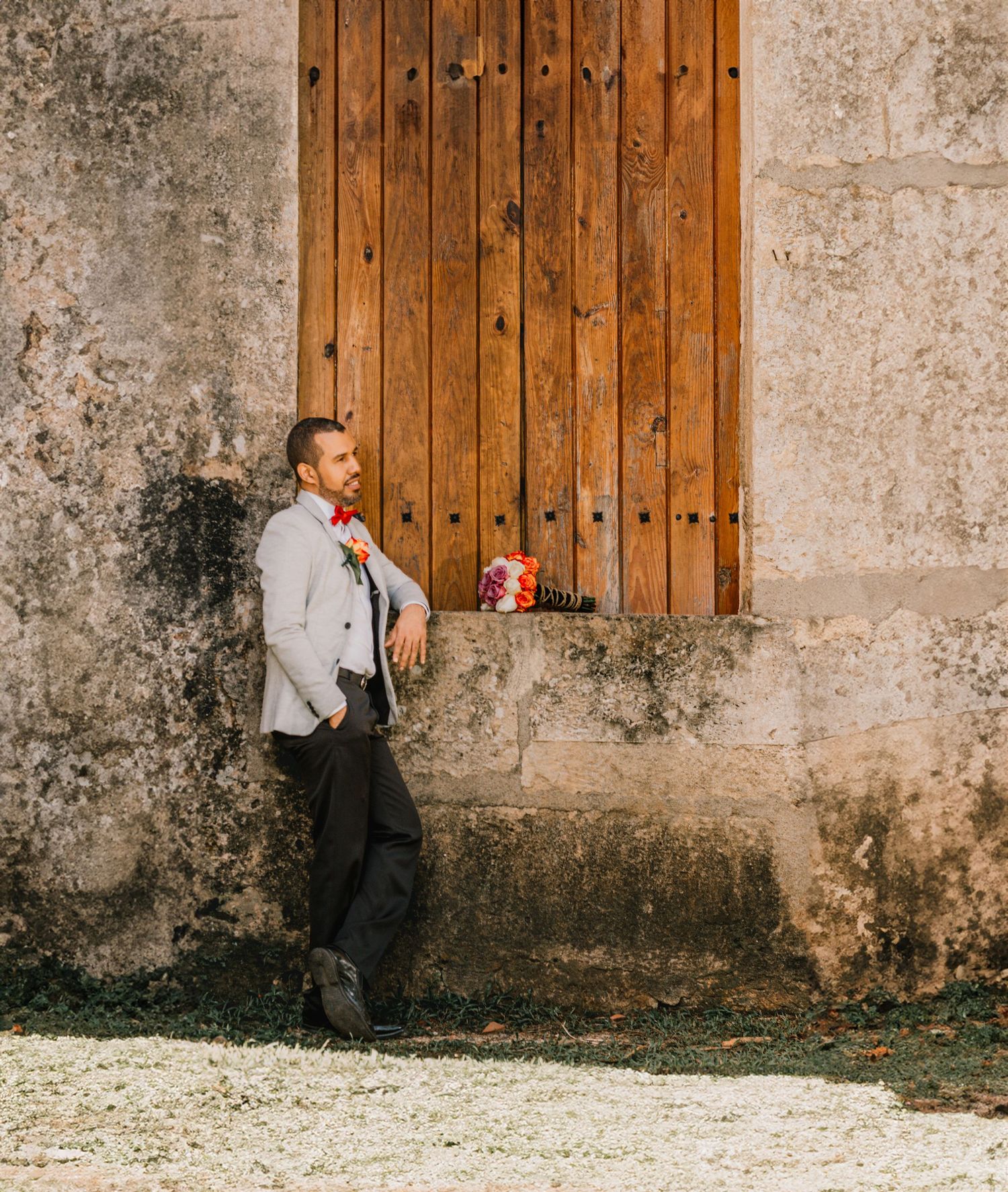 Groom in grey blazer leaning against rustic wooden door at stone building.