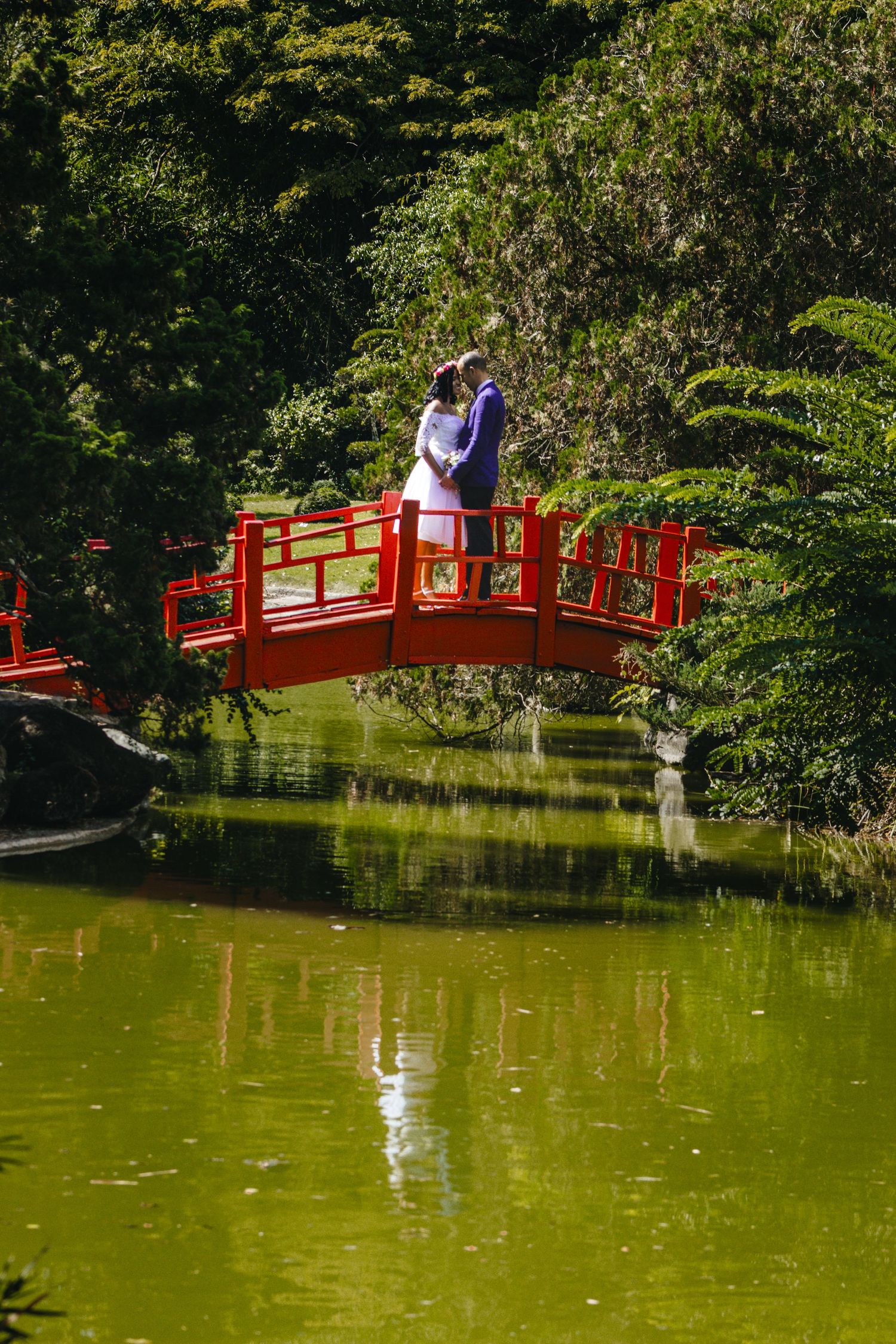 Newlywed couple sharing a romantic moment on a red arched bridge over a serene green pond in a Japanese garden.