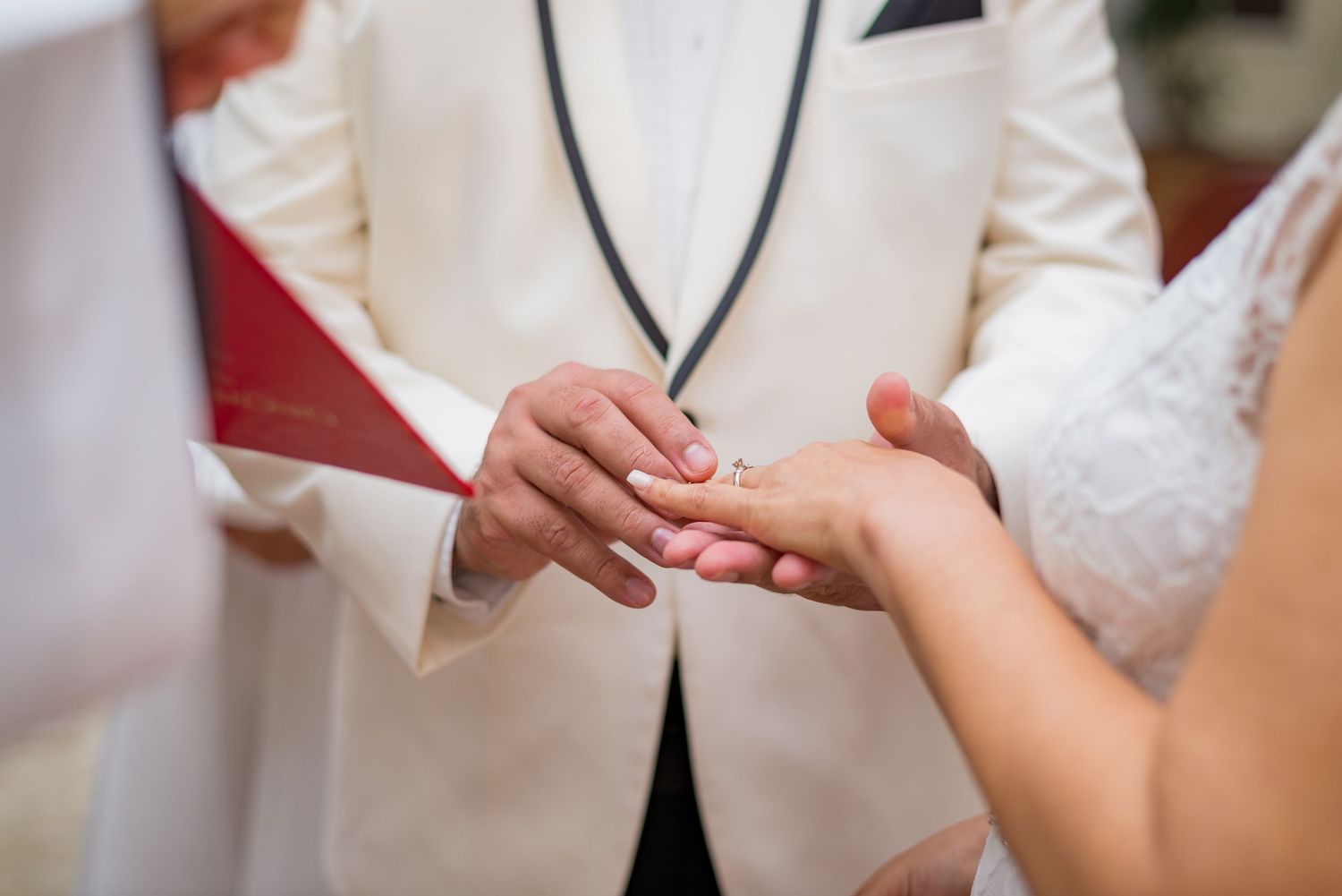Groom in white tuxedo placing wedding ring on brides finger during ceremony.