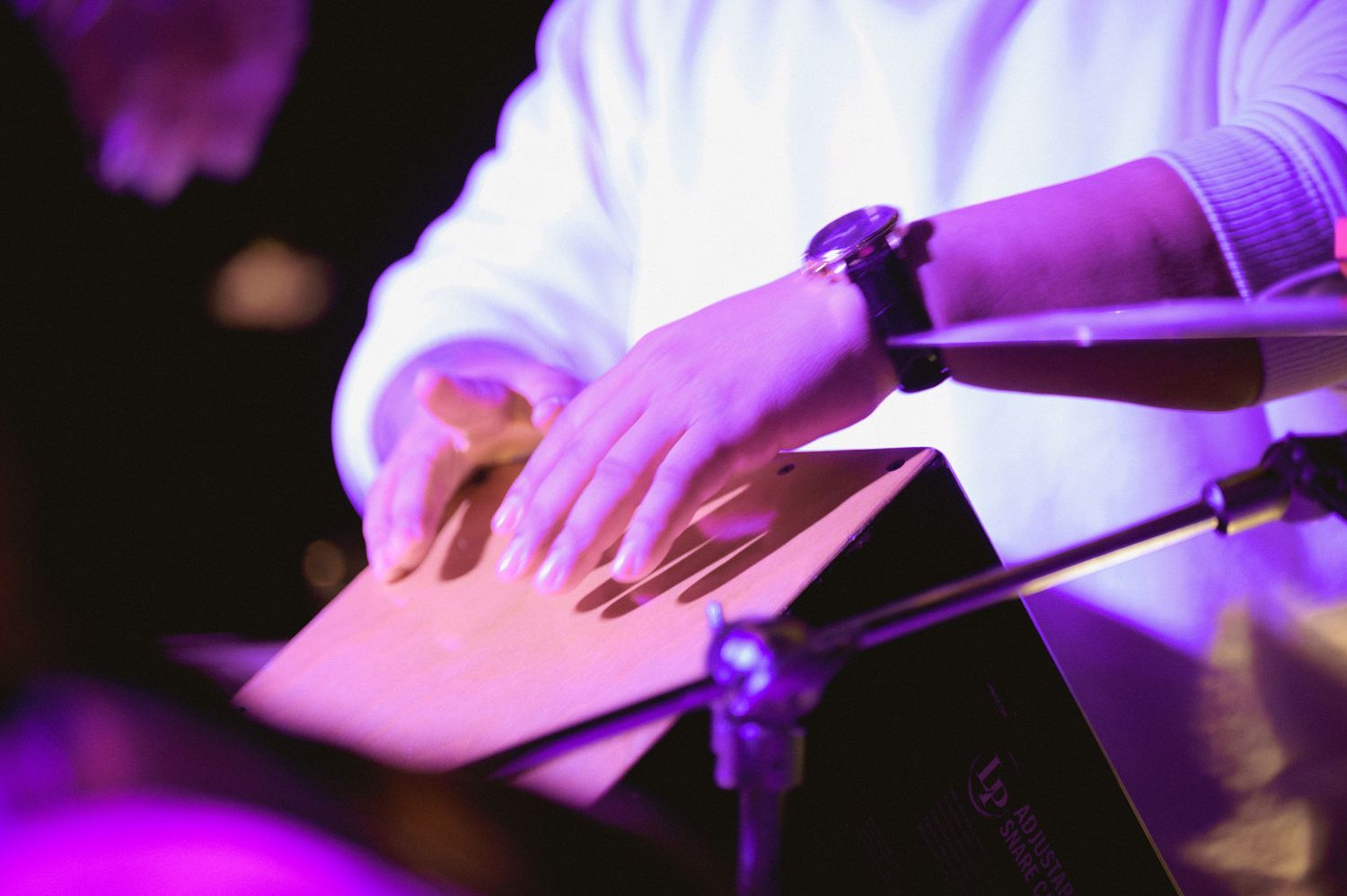A musician in a white shirt plays cymbals under purple stage lighting during a live performance.
