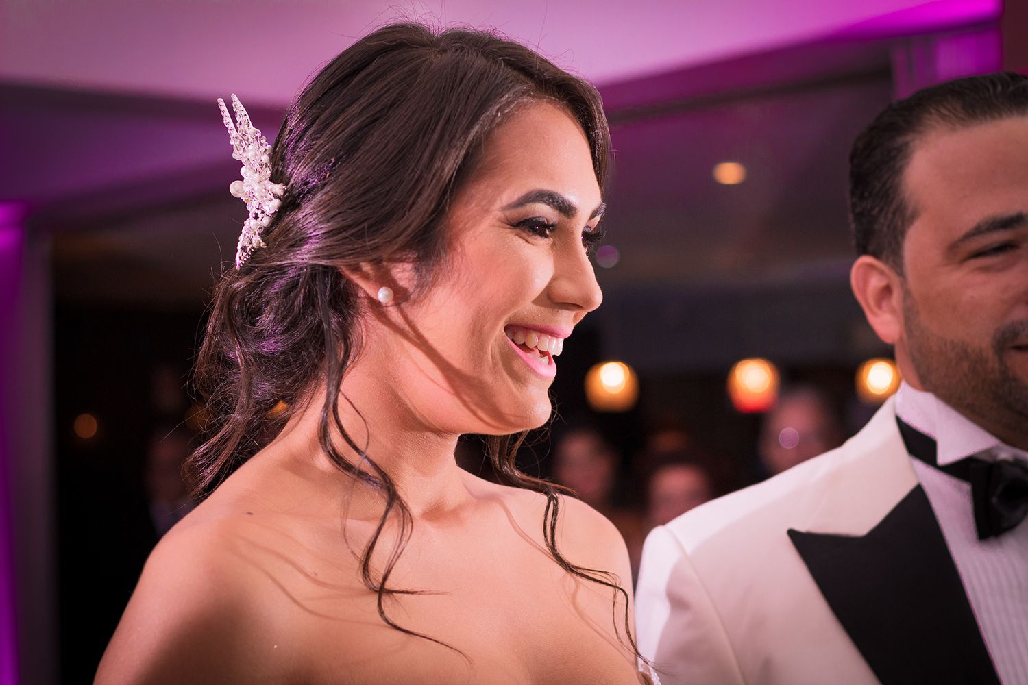 Radiant bride smiling joyfully in strapless lace gown with floral hair accessory at wedding.