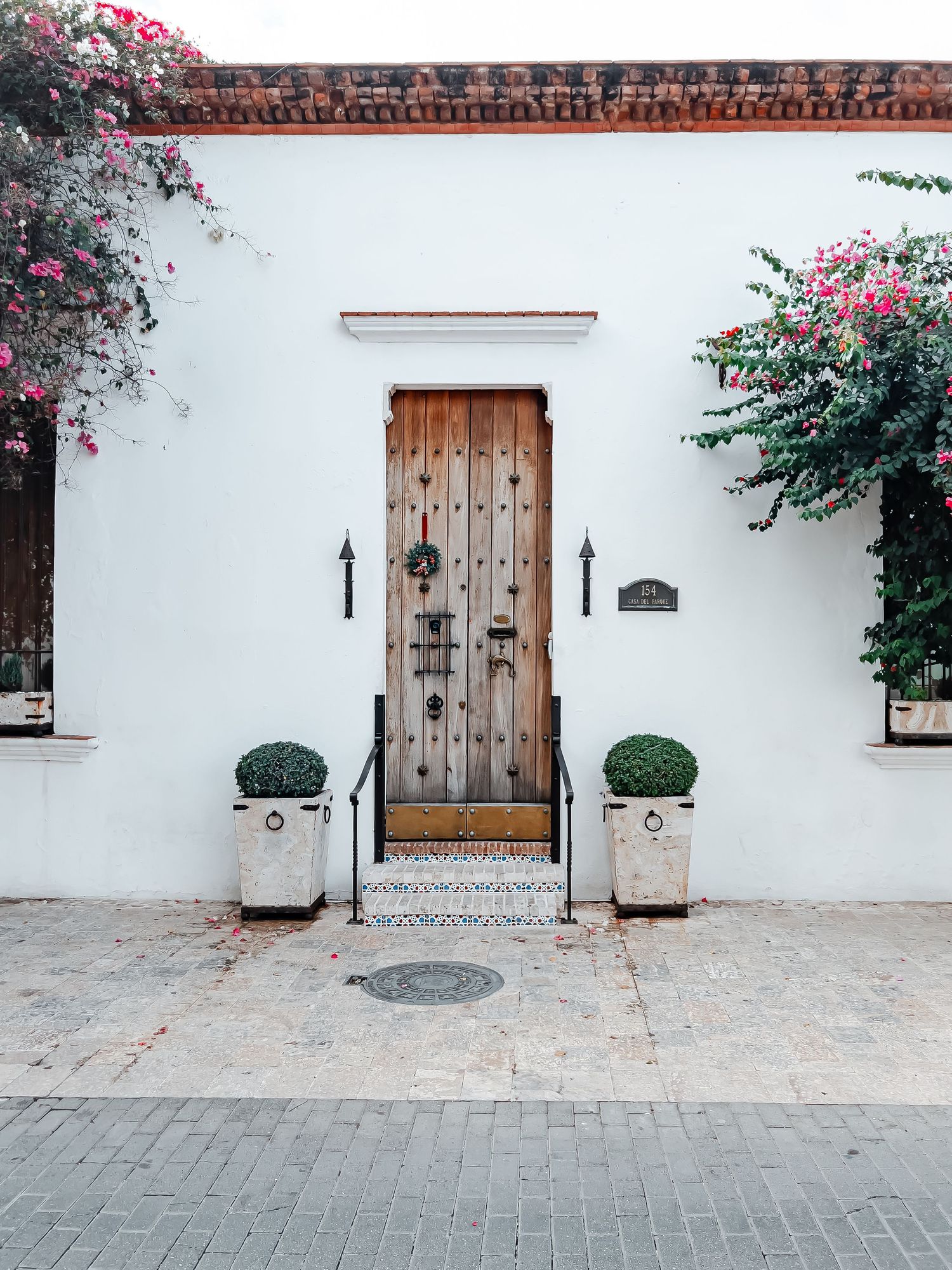 White Spanish colonial building facade with ornate wooden door flanked by topiary plants and blooming bougainvillea.