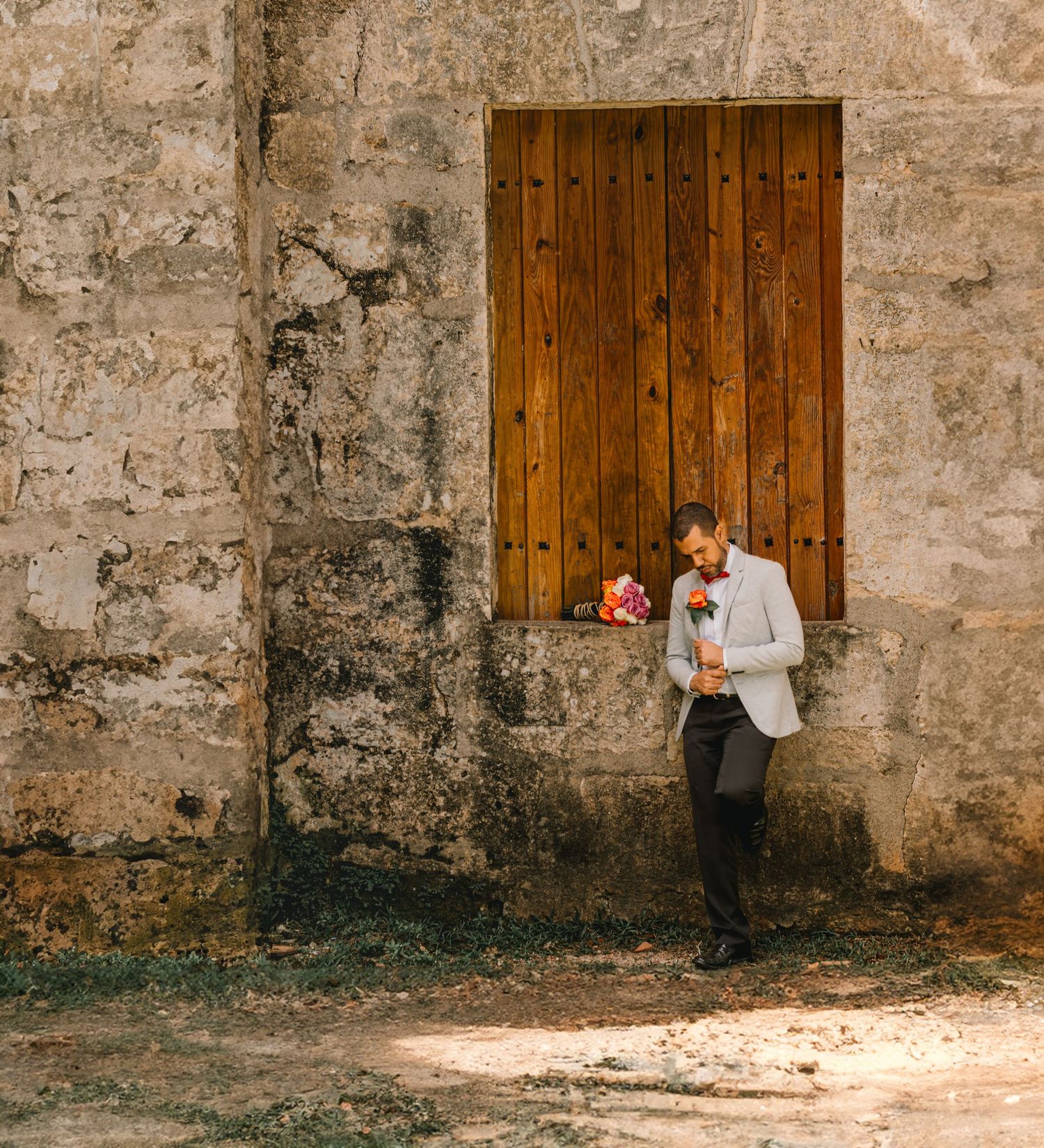 Groom in grey blazer leaning against rustic stone wall holding colorful bouquet.