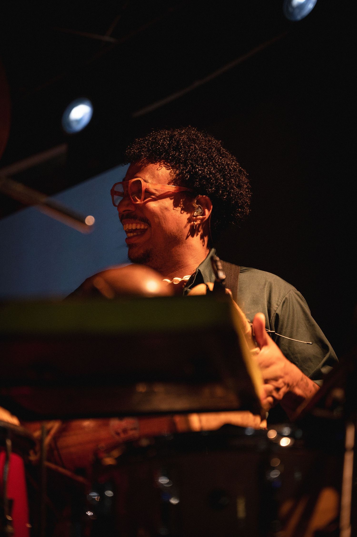 A smiling drummer with curly hair and pink glasses playing drums on a warmly lit stage.