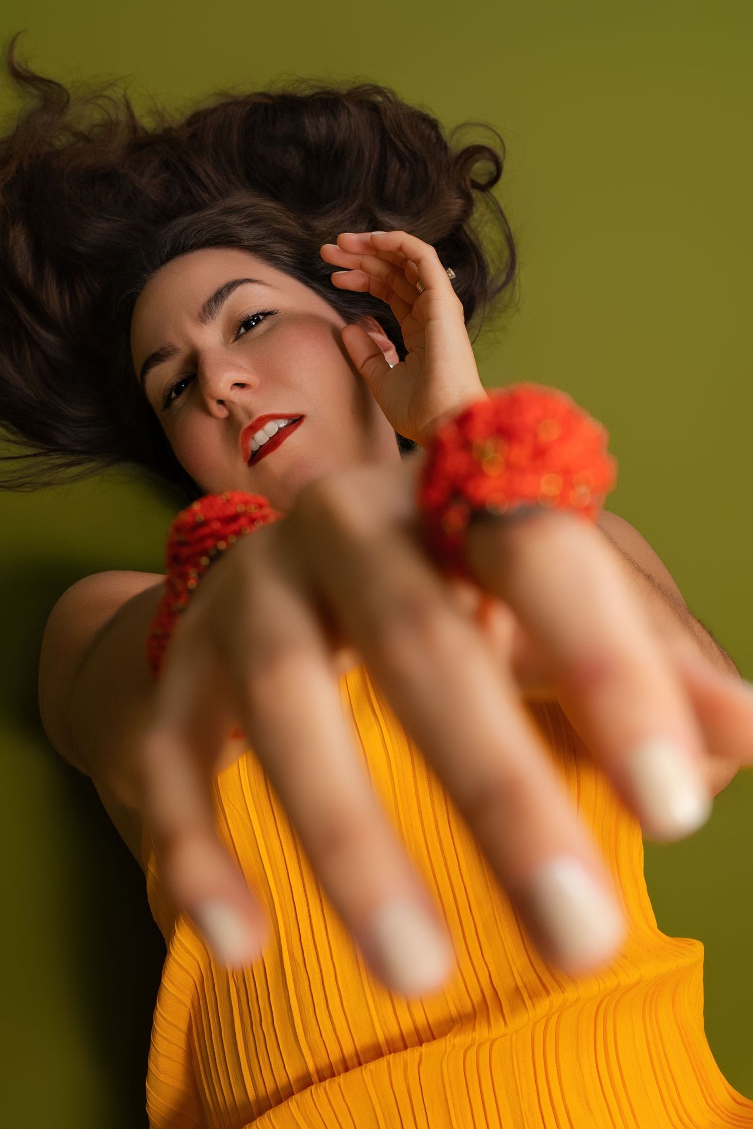 Woman in yellow dress lying down on green background pointing at camera wearing orange floral bracelets and necklace.