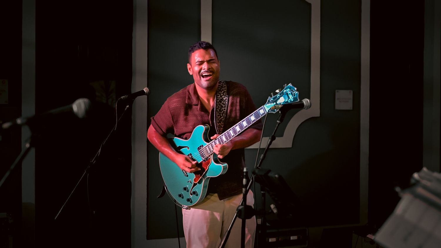 A smiling male guitarist plays a teal semi-hollow electric guitar on a dimly lit stage.