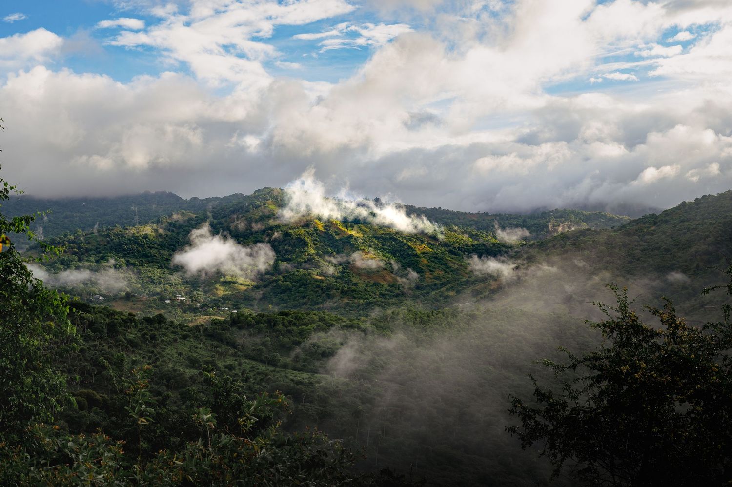 Misty green mountains covered in lush tropical forest with low clouds rolling through the valleys under a partly cloudy sky.