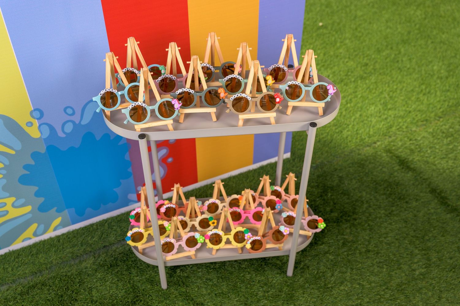 Two-tier wooden display stand holding colorful party favor sunglasses on green grass background.