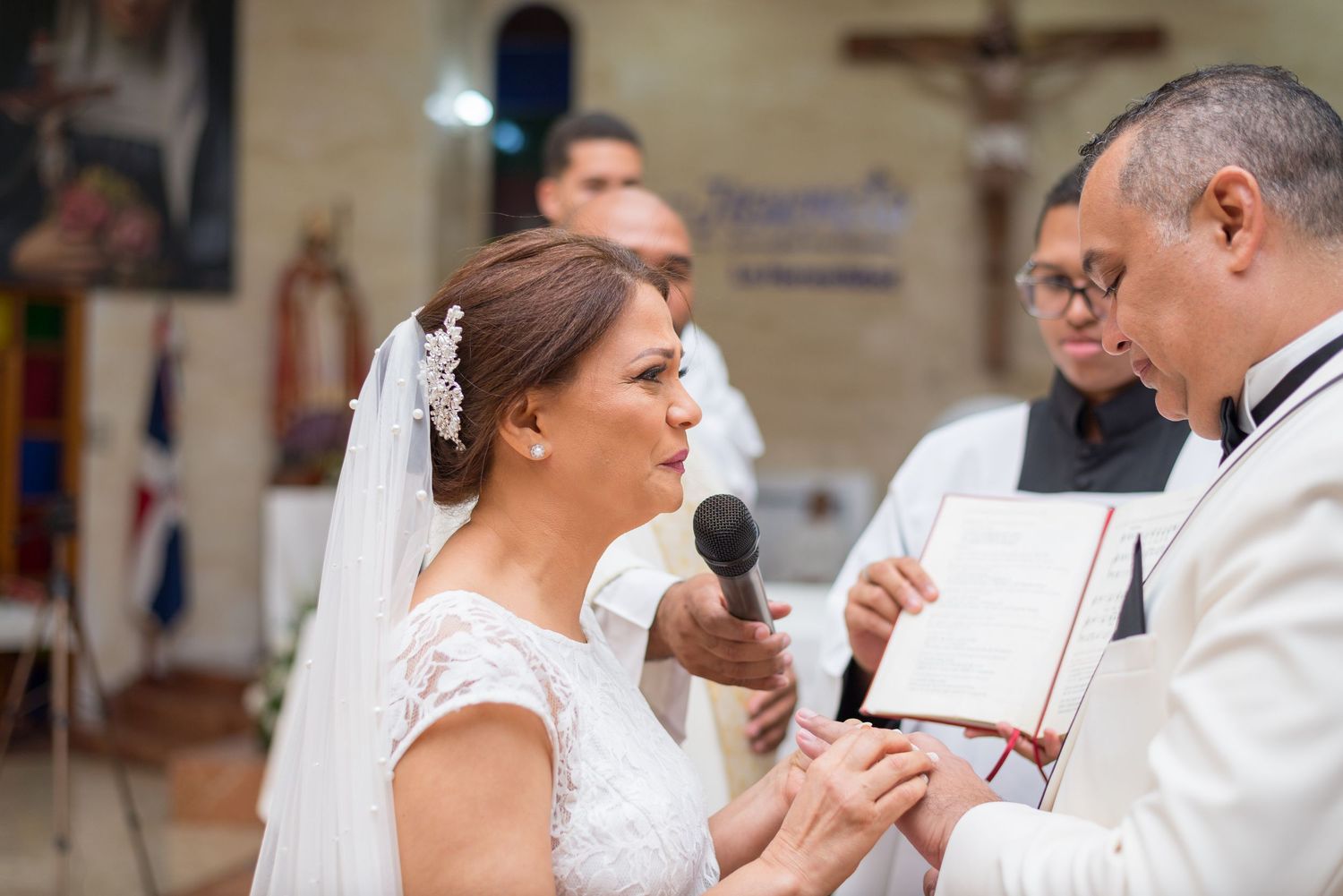 Bride in white veil speaking vows into microphone at Catholic church wedding ceremony.