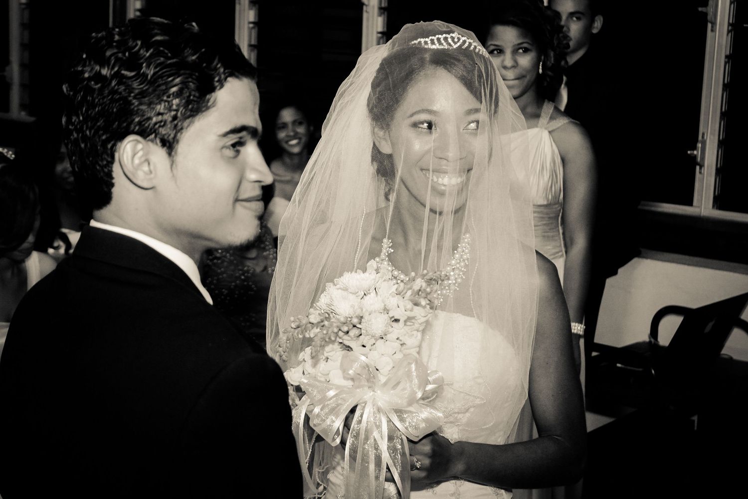 Black and white photo of smiling bride in veil and tiara with groom at wedding ceremony.