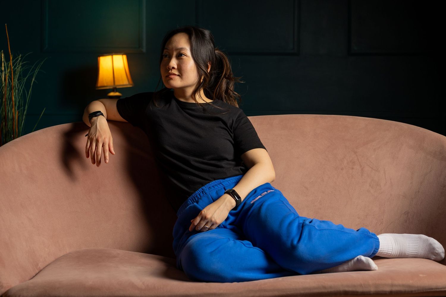 A relaxed young adult Asian woman sitting in casual loungewear on a pink couch in front of a deep green wall. It's a flash studio portrait.