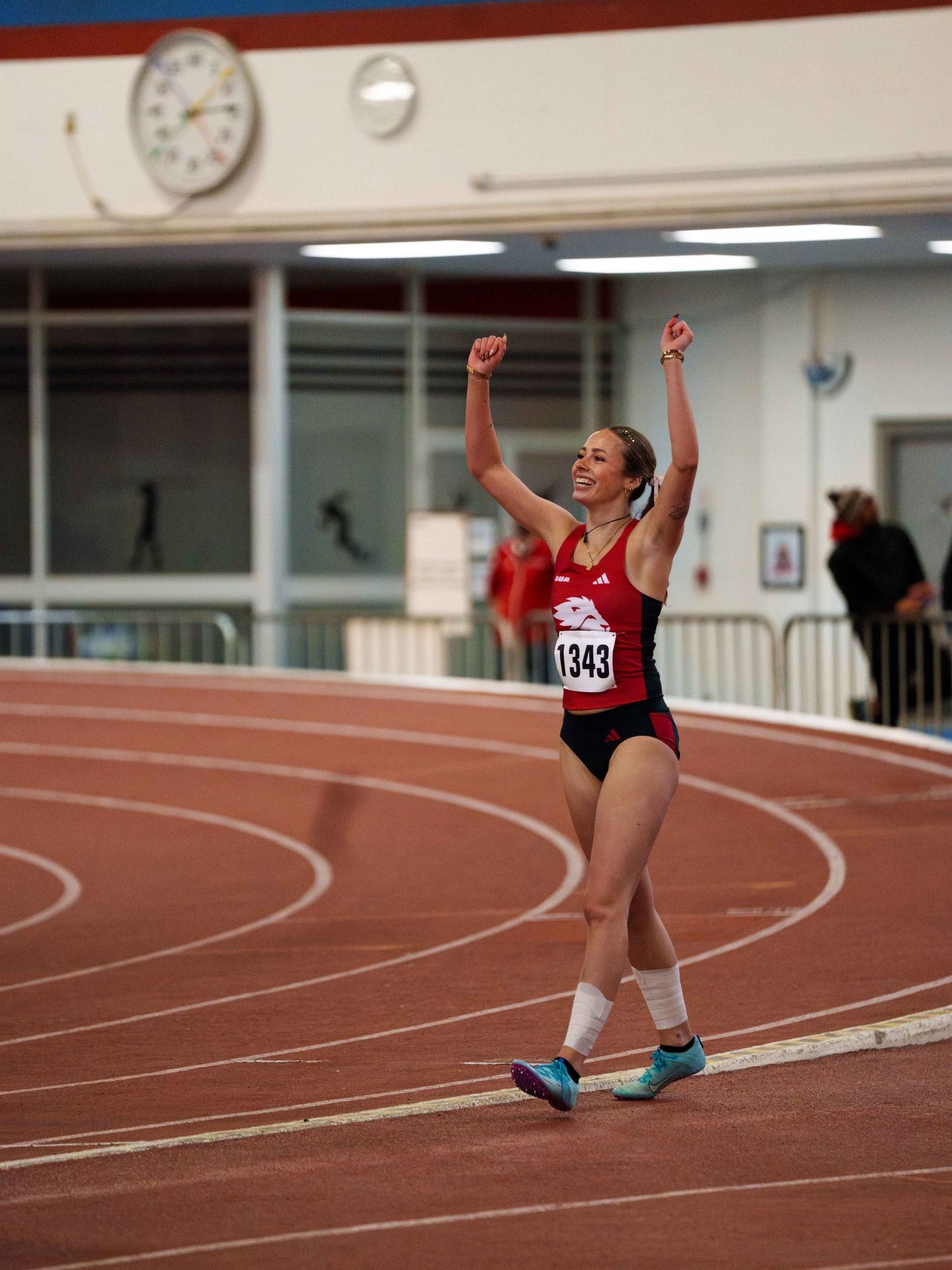 A young white female track runner smiles and waves at people in the stands as she walks back after completing her race.