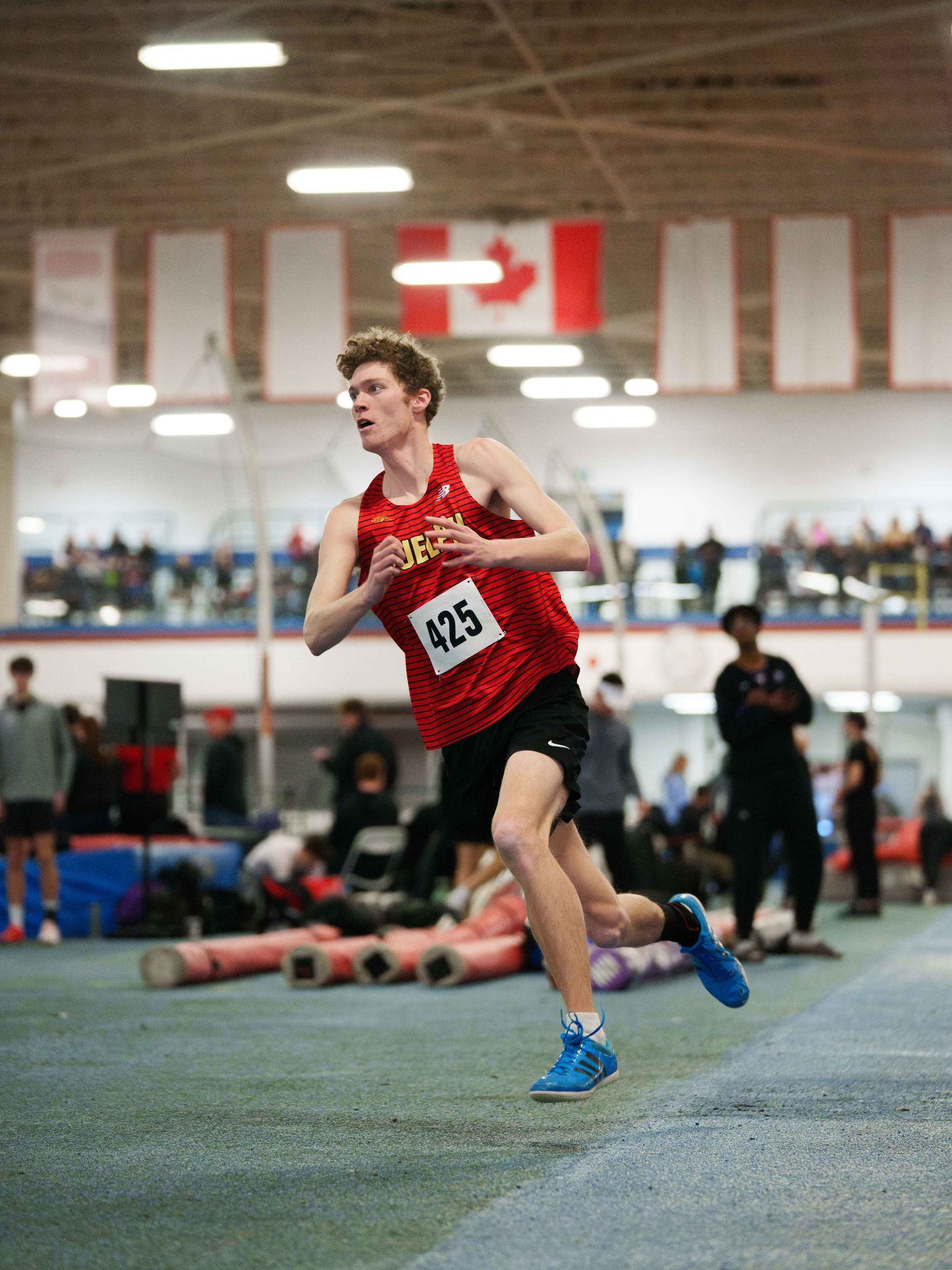 A young white male high jumper frozen mid starting run, illuminated by a line of gymnasium lights above him. The Canadian flag in the back.