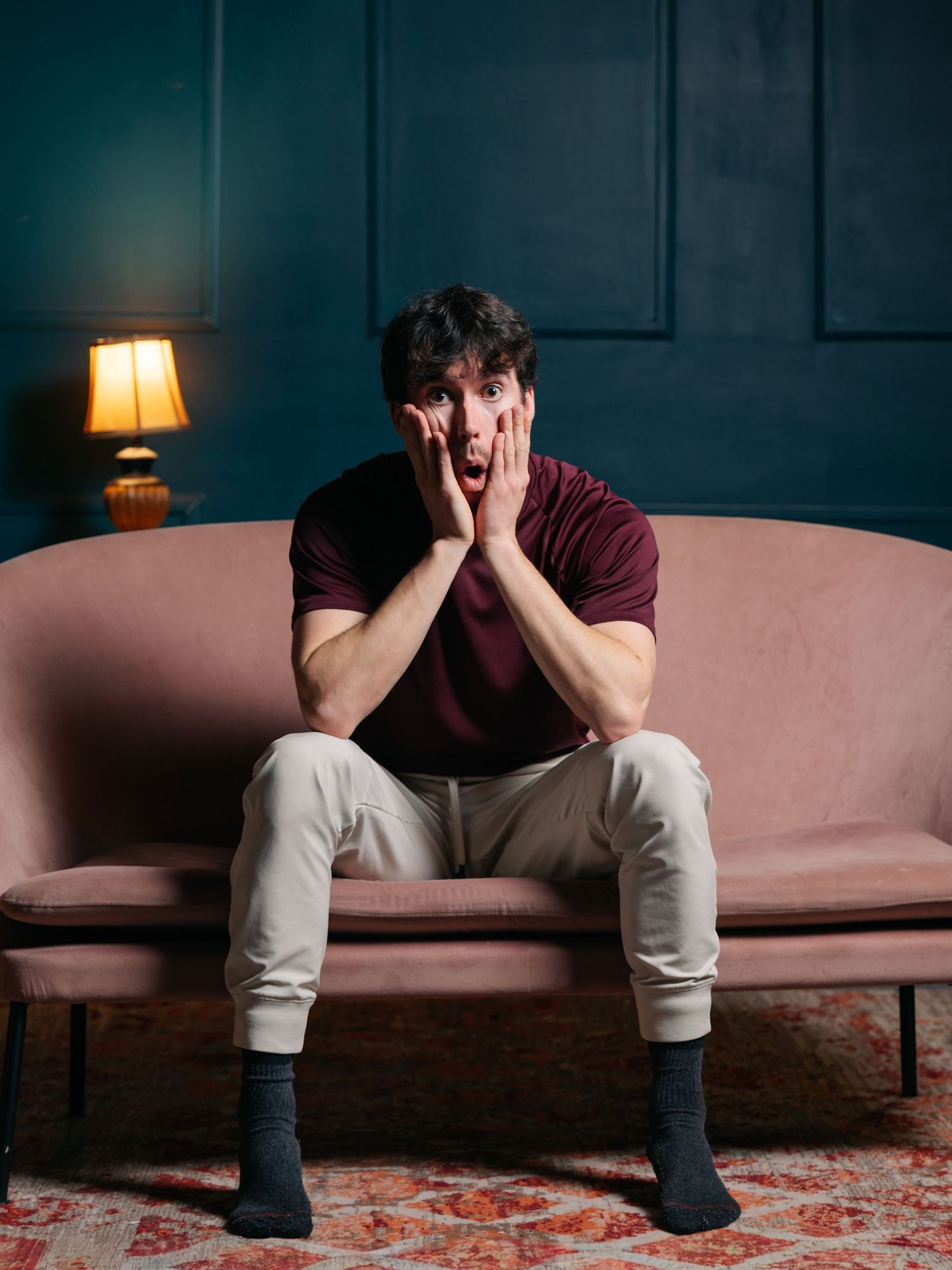Flash studio portrait of a young white man making a surprised face sitting on a pink couch in front of a deep green wall.