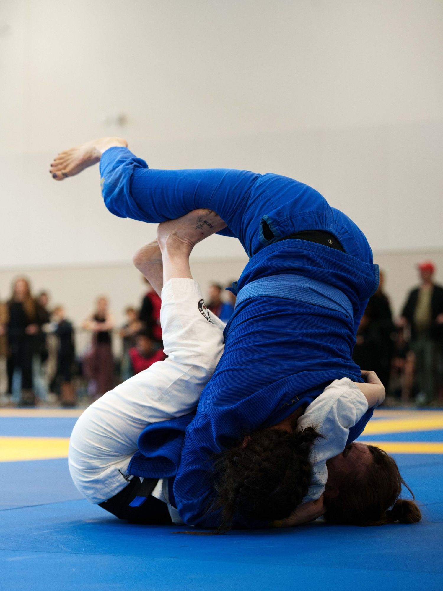 Two female BJJ fighters - one in a blue gi and the other in a white gi - grapple on the ground.