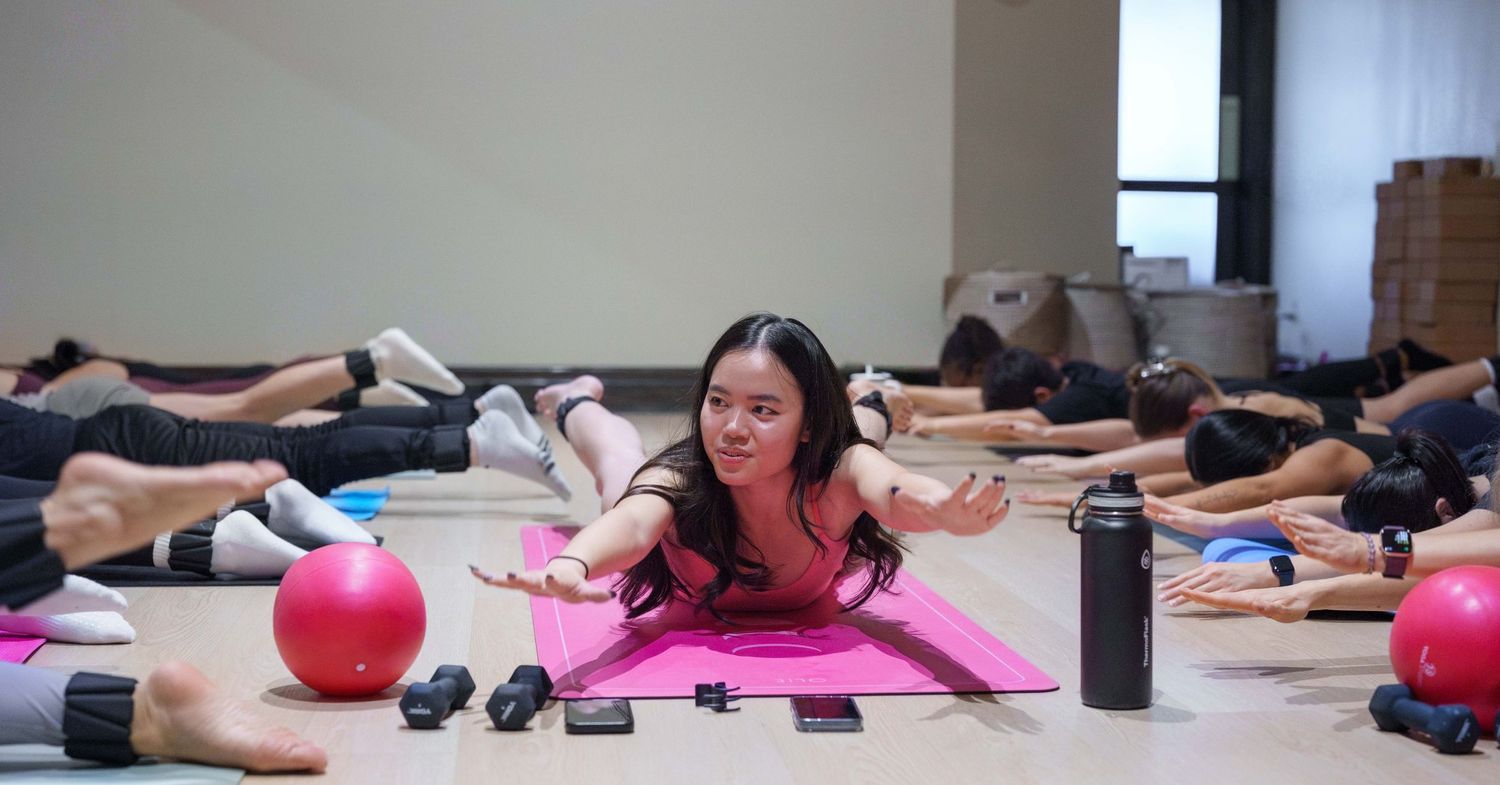 A young Asian woman teaching pilates wearing a matching pink set atop a pink mat with a deep pink ball next to her. Students are visible.