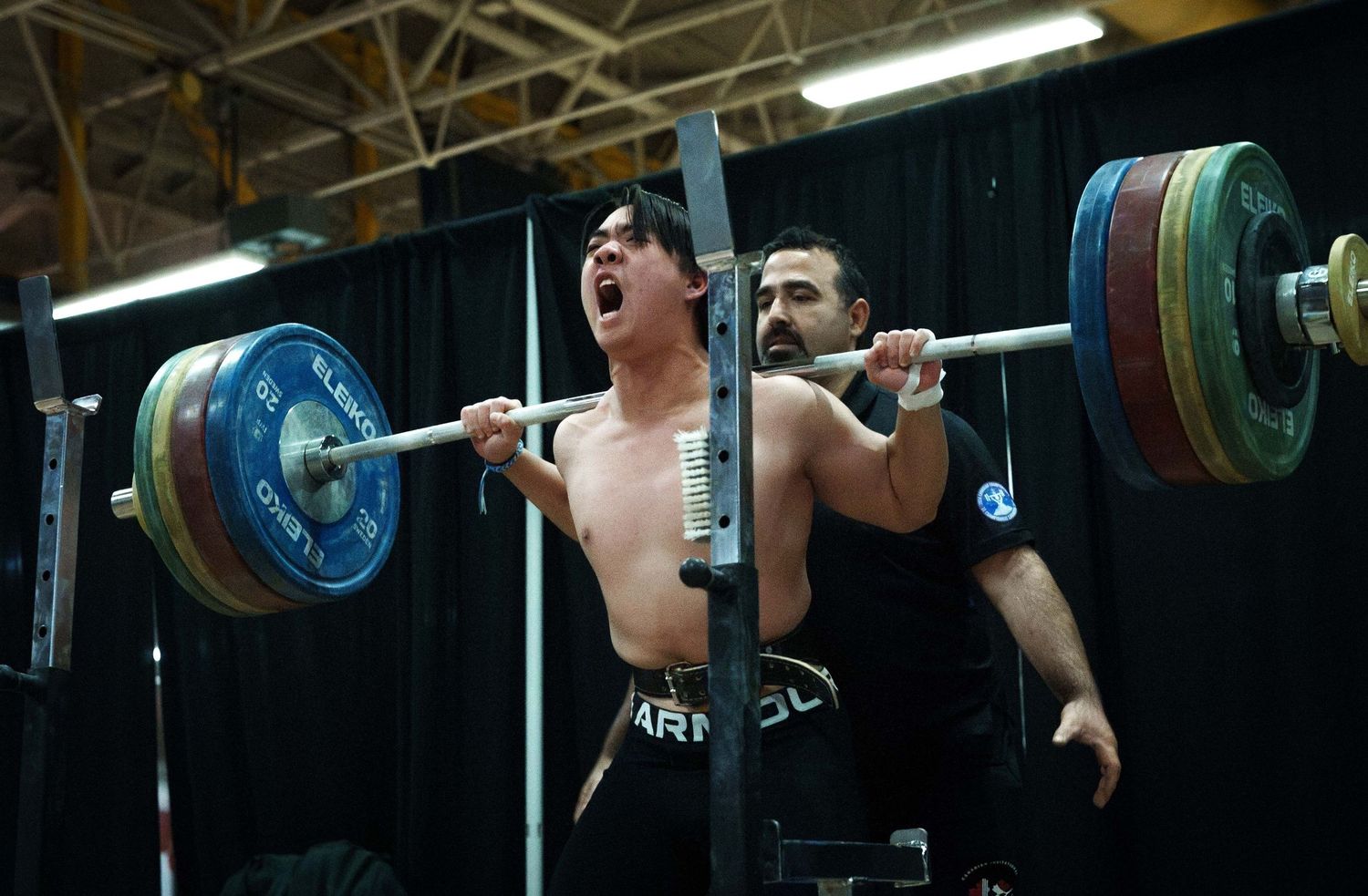 Young, Asian male weightlifter shouts as he successfully completes a PR attempt for his back squat. He is not wearing a shirt.