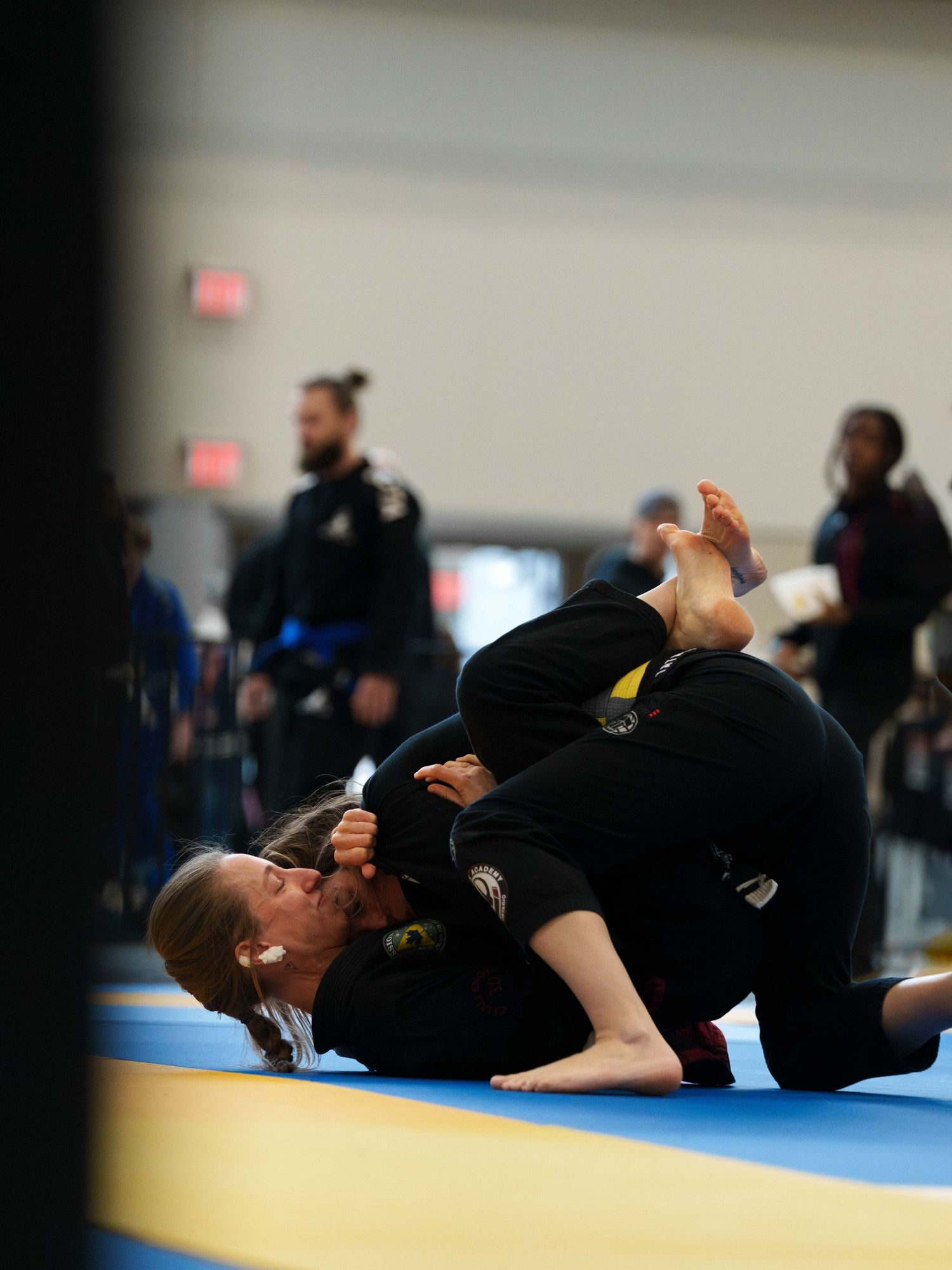 Two female BJJ fighters in black gis grapple on the ground.